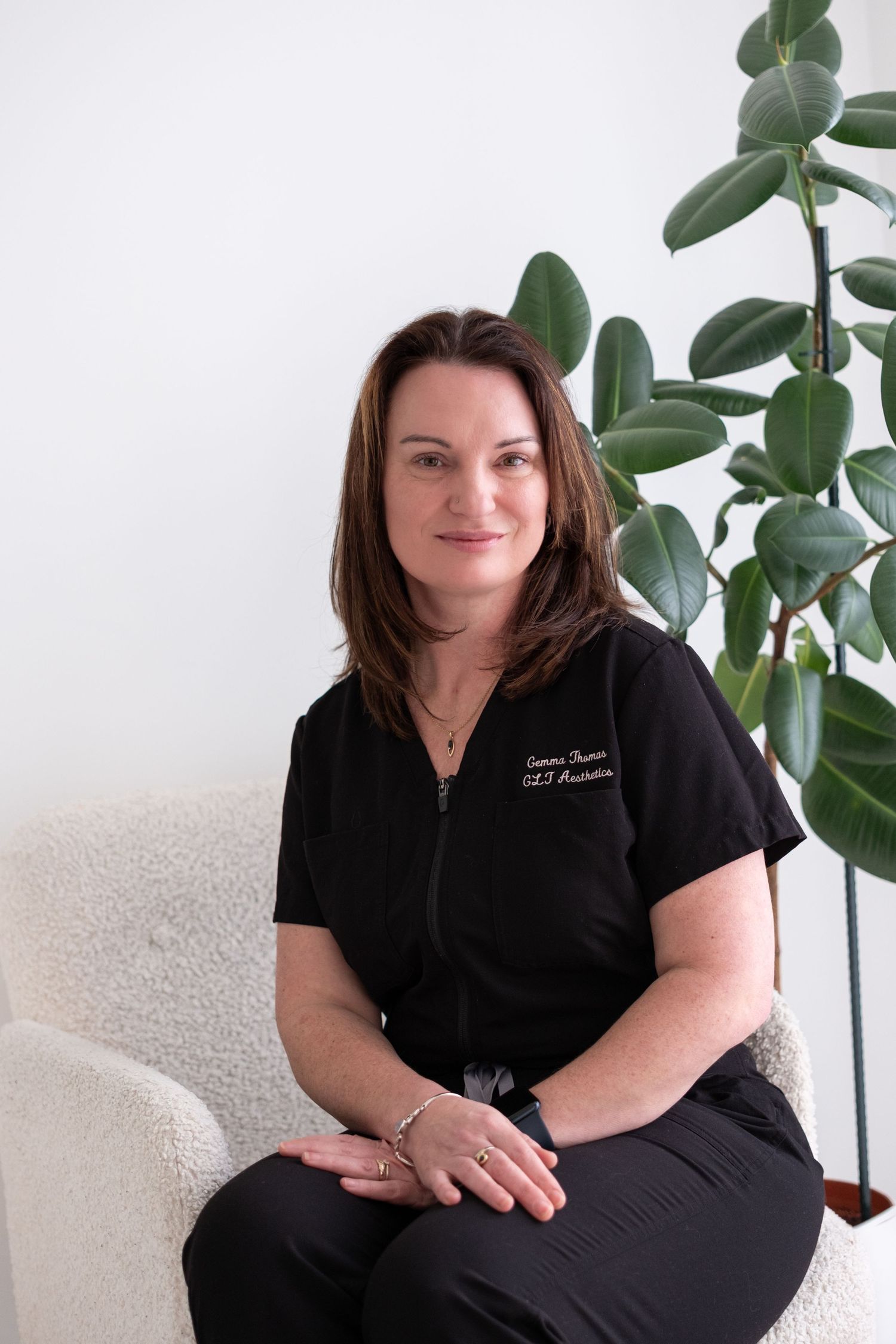 Woman in black medical scrubs sits in white chair beside rubber plant in bright clinical setting.