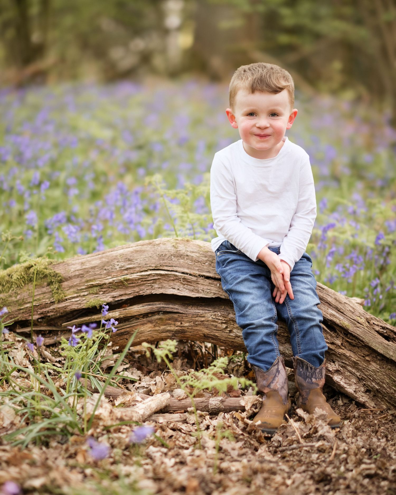 Young boy in white shirt and jeans sits smiling on a log surrounded by purple bluebells in a woodland setting.