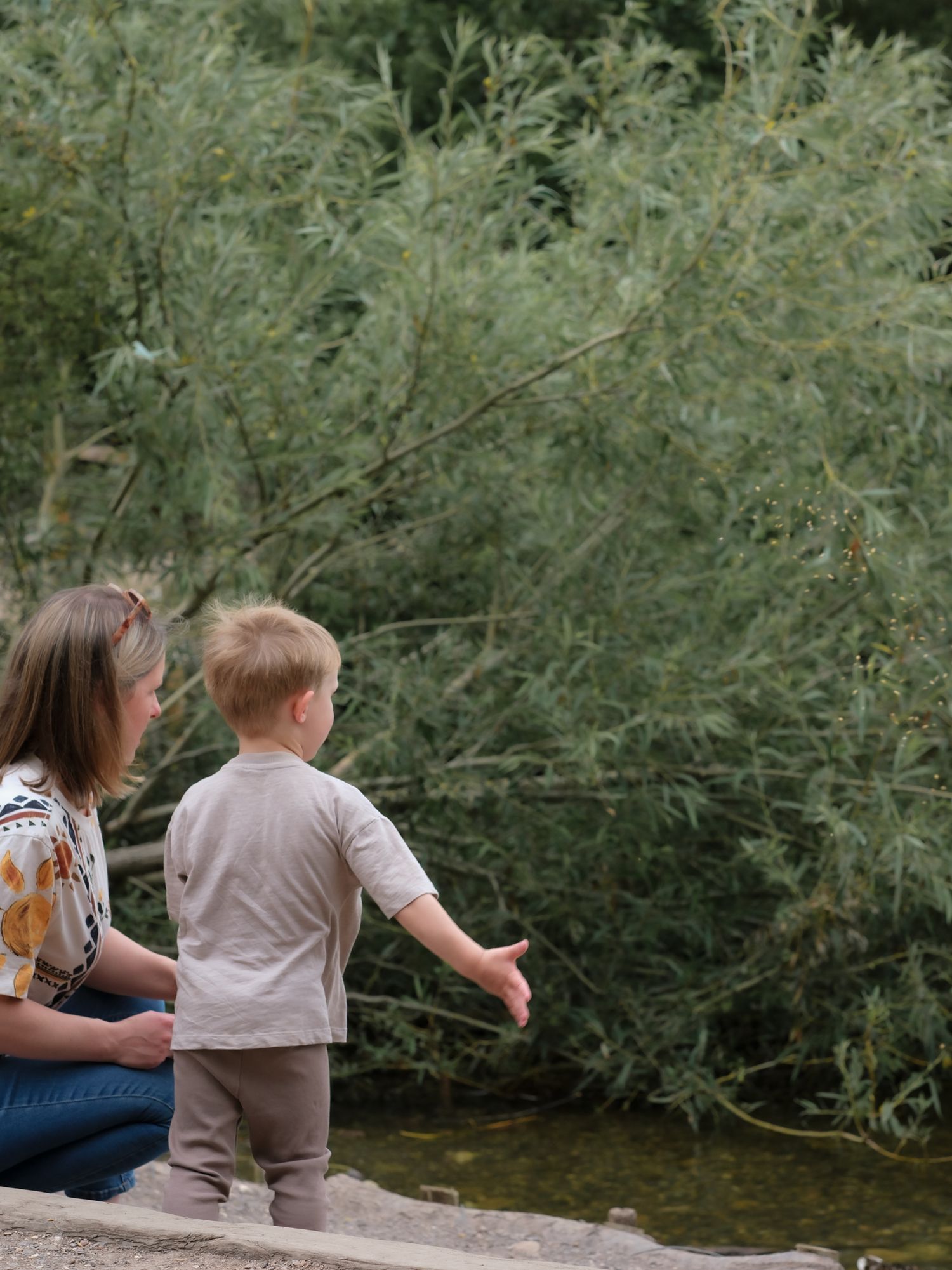 A mother and toddler boy explore a lush green park near a pond, the child reaching out curiously.