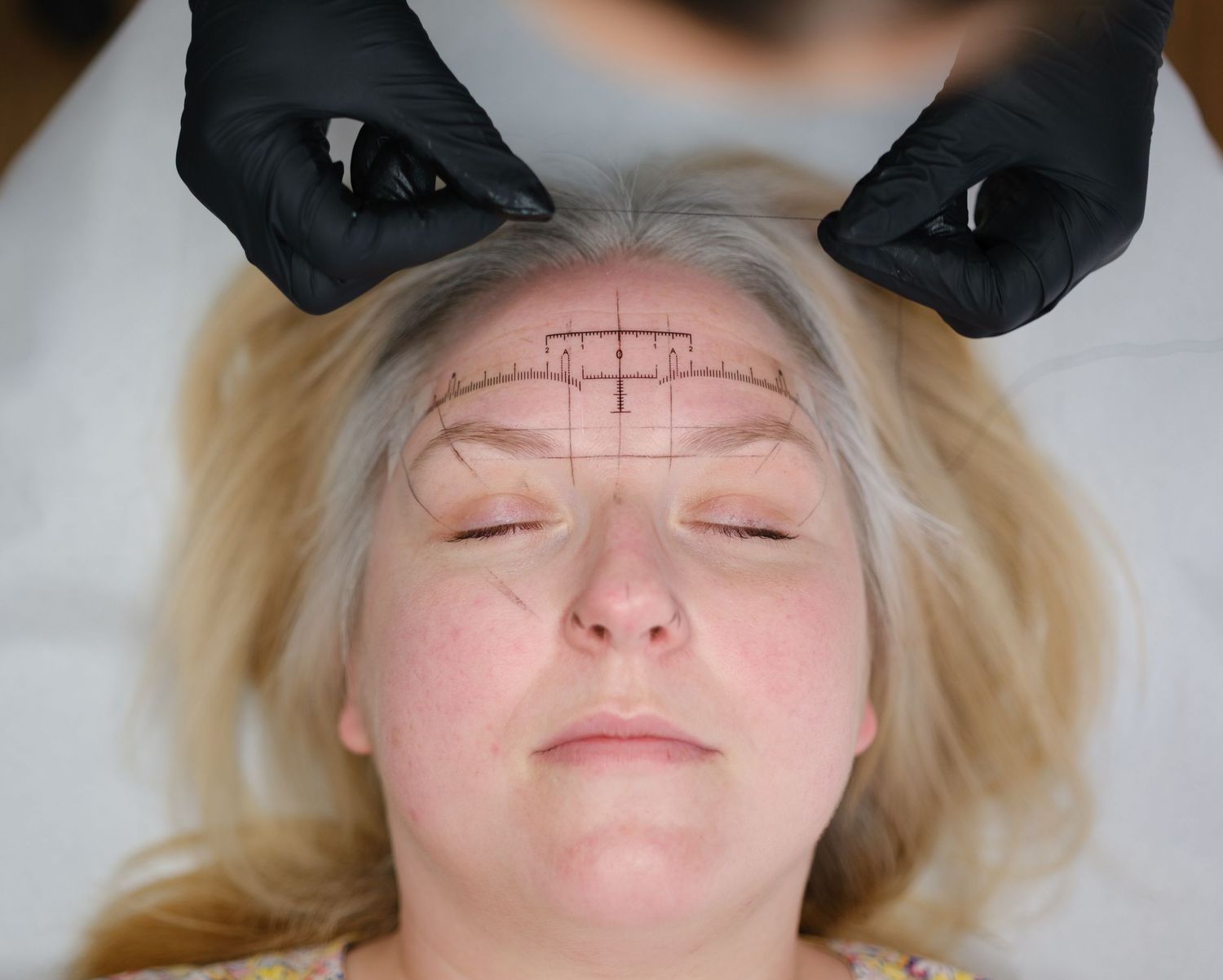 Black gloved hands measuring and mapping eyebrow symmetry with guidelines drawn on a blonde womans forehead.