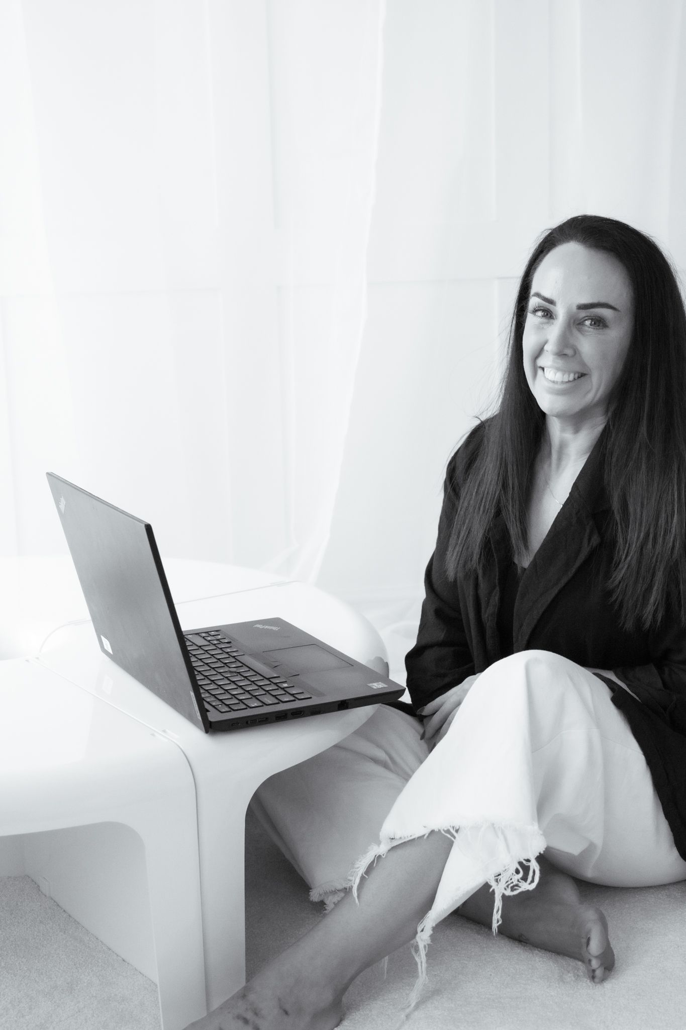 Smiling woman in black jacket sits cross-legged with laptop in bright white room, monochrome photo.