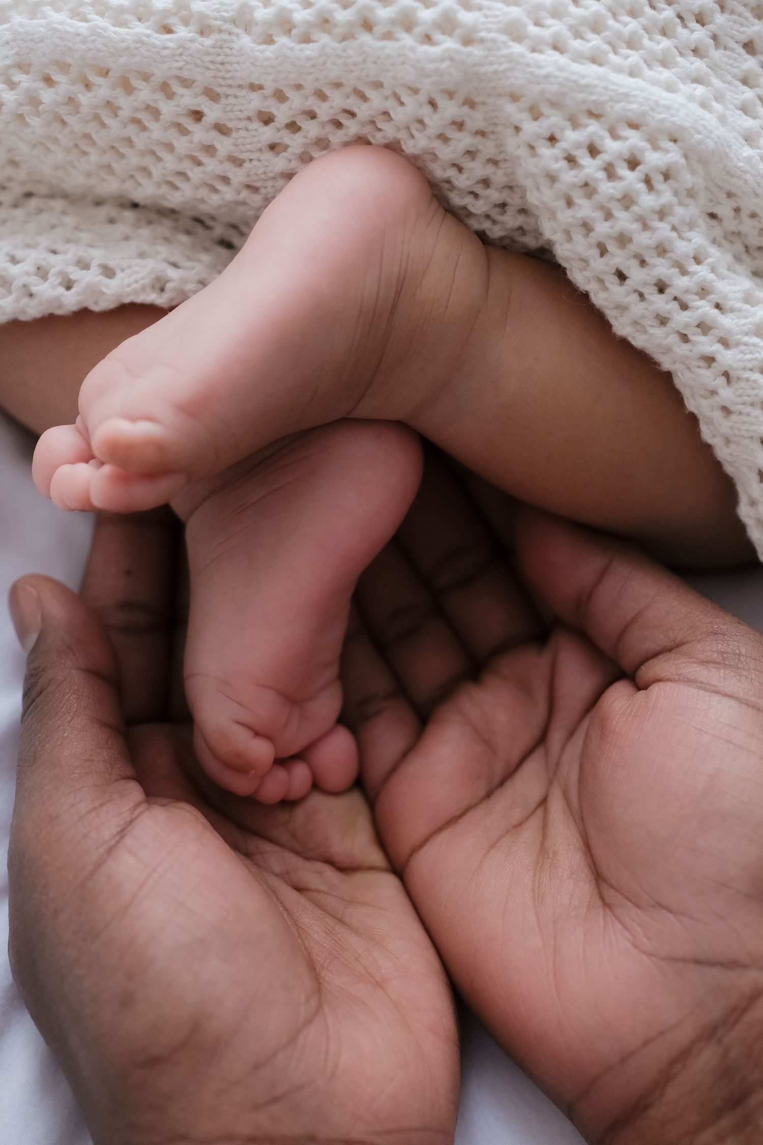 Parent hands cradling tiny newborn baby feet under white knit blanket.