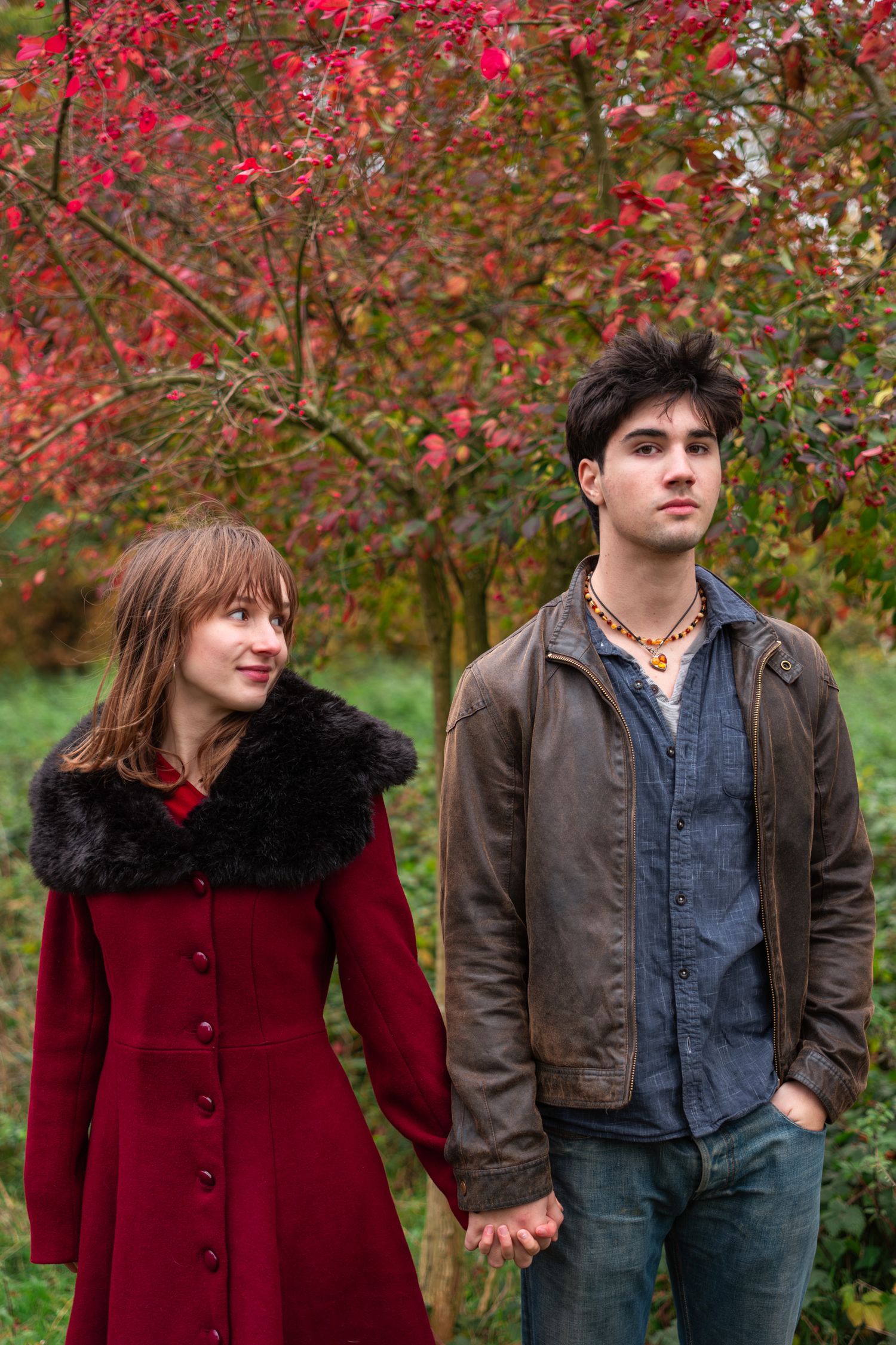 Young couple holding hands standing together in autumn park with vibrant red foliage background.