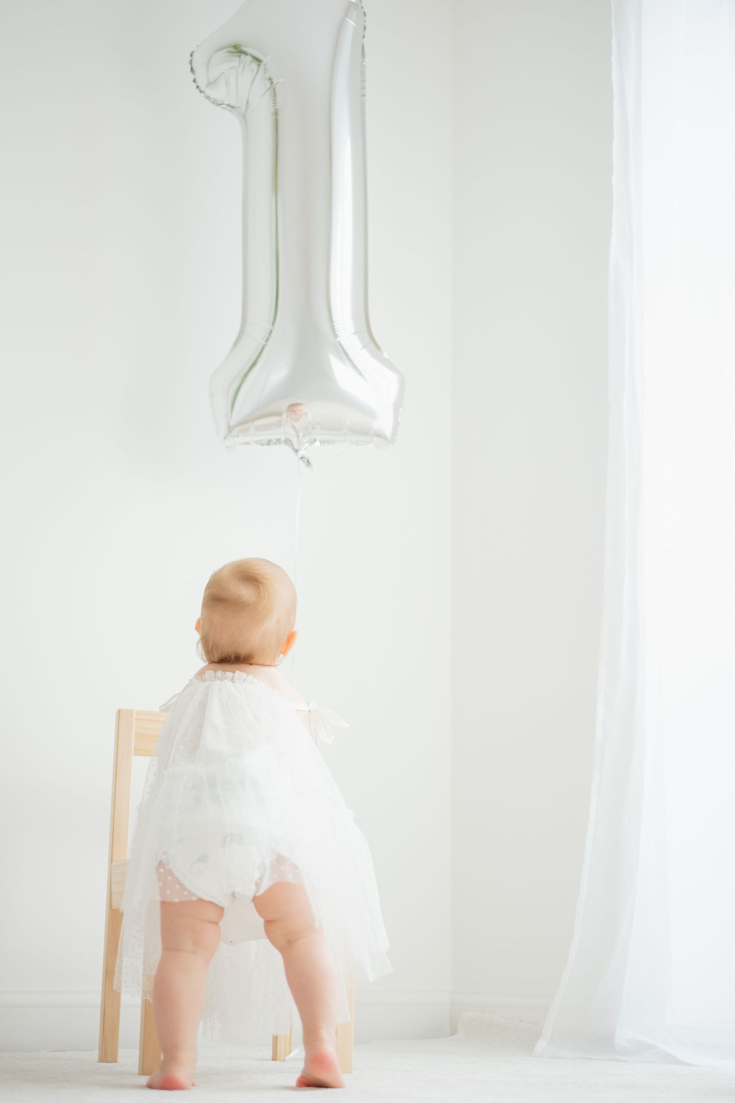 Baby in white dress standing at chair with silver number 1 balloon for first birthday.