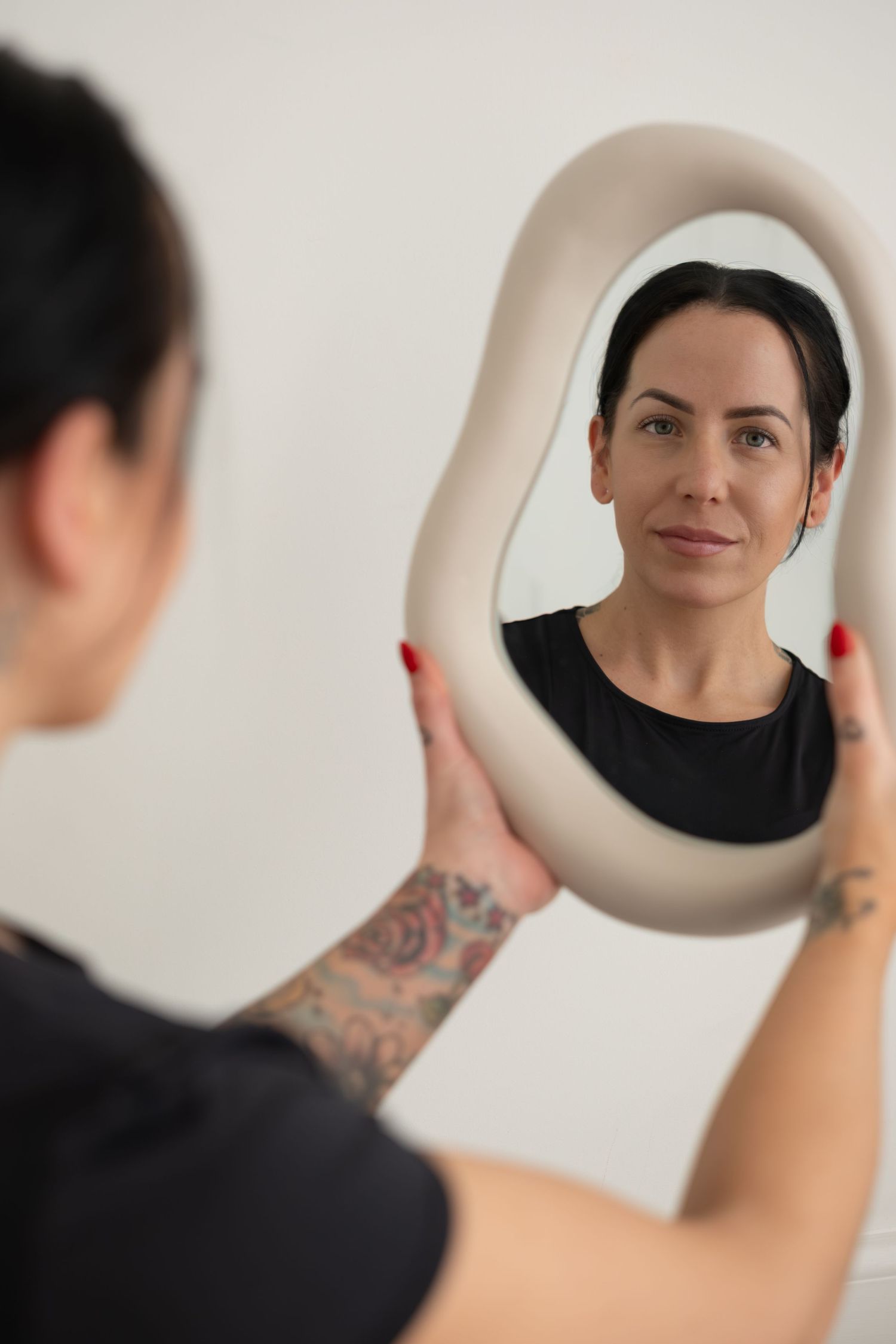 Tattooed woman with red nails holds wavy mirror reflecting her face against white background.
