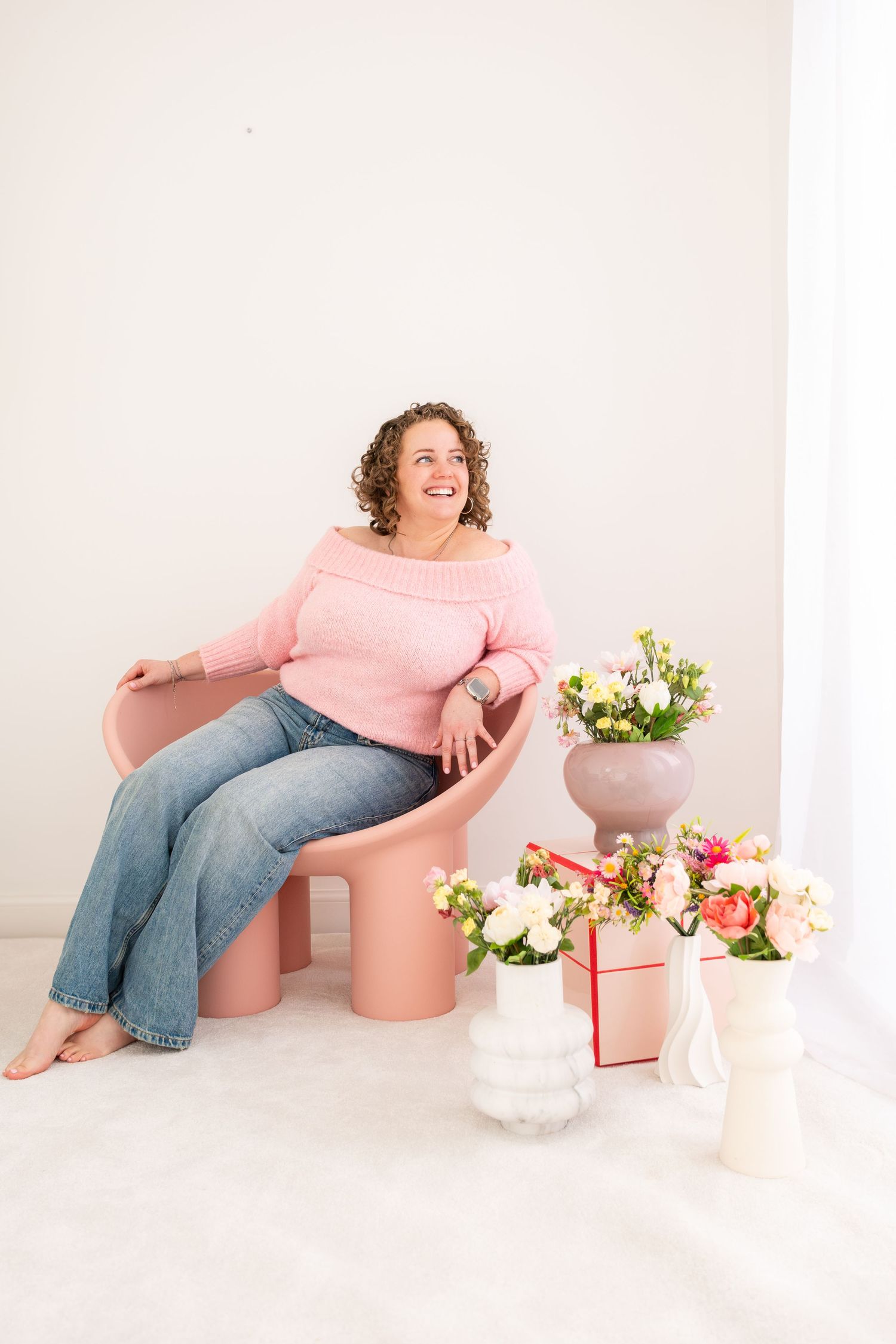 Woman in pink sweater and jeans sitting in pink chair surrounded by floral arrangements in bright white studio.