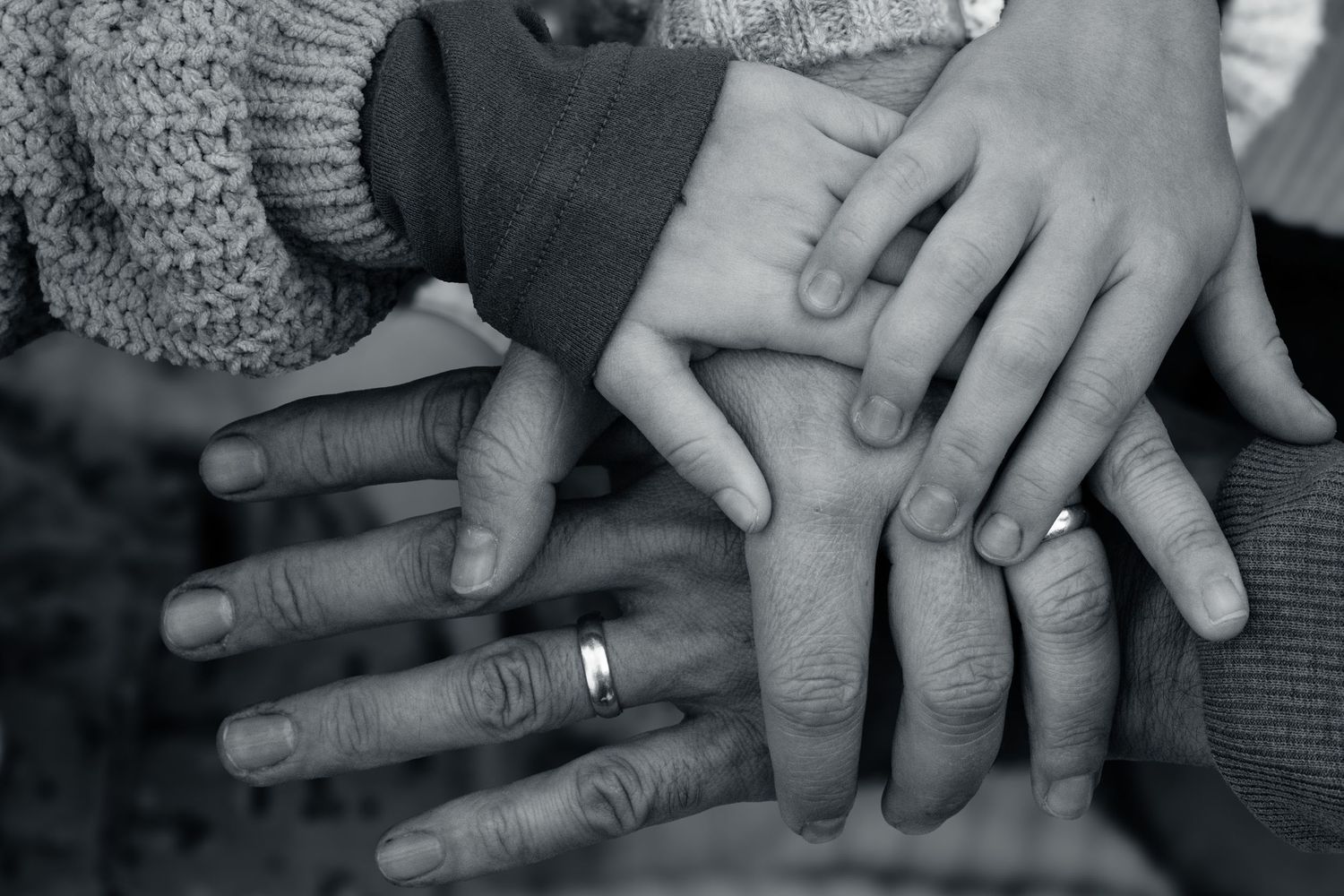 Black and white close-up of multiple family hands stacked together showing wedding ring.