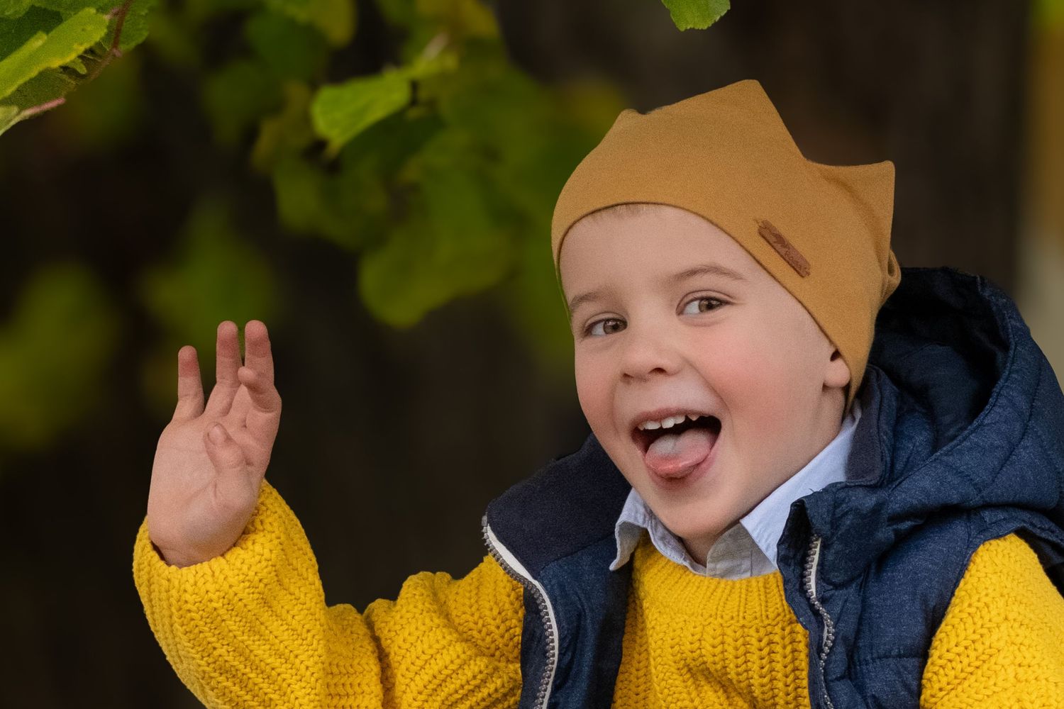 Playful young boy in yellow sweater and brown beanie sticks out tongue and waves hand in autumn park.