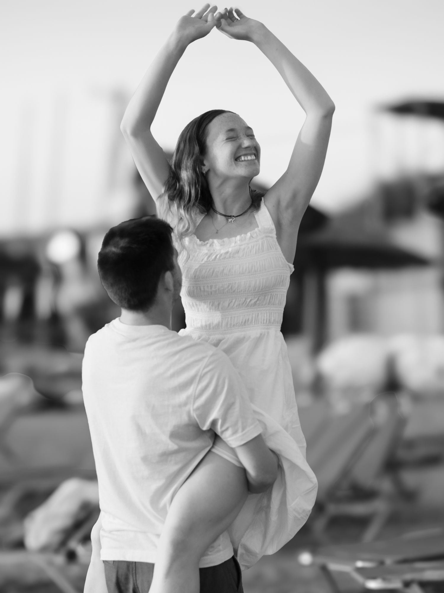 Black and white photo of a joyful woman in white dress being lifted by a man on a sunny beach.