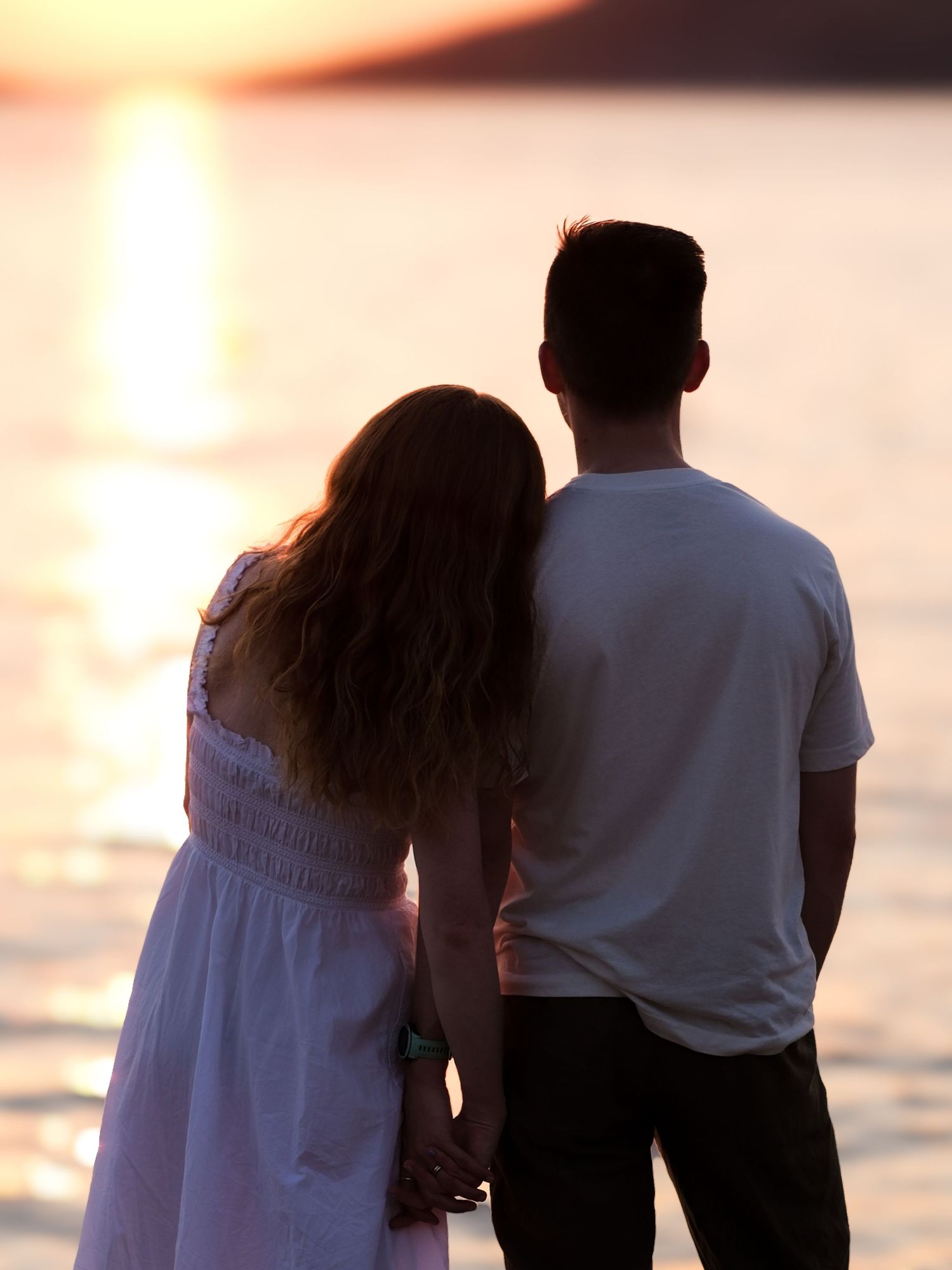 A romantic couple holds hands while watching a golden sunset over calm ocean waters from the shore.
