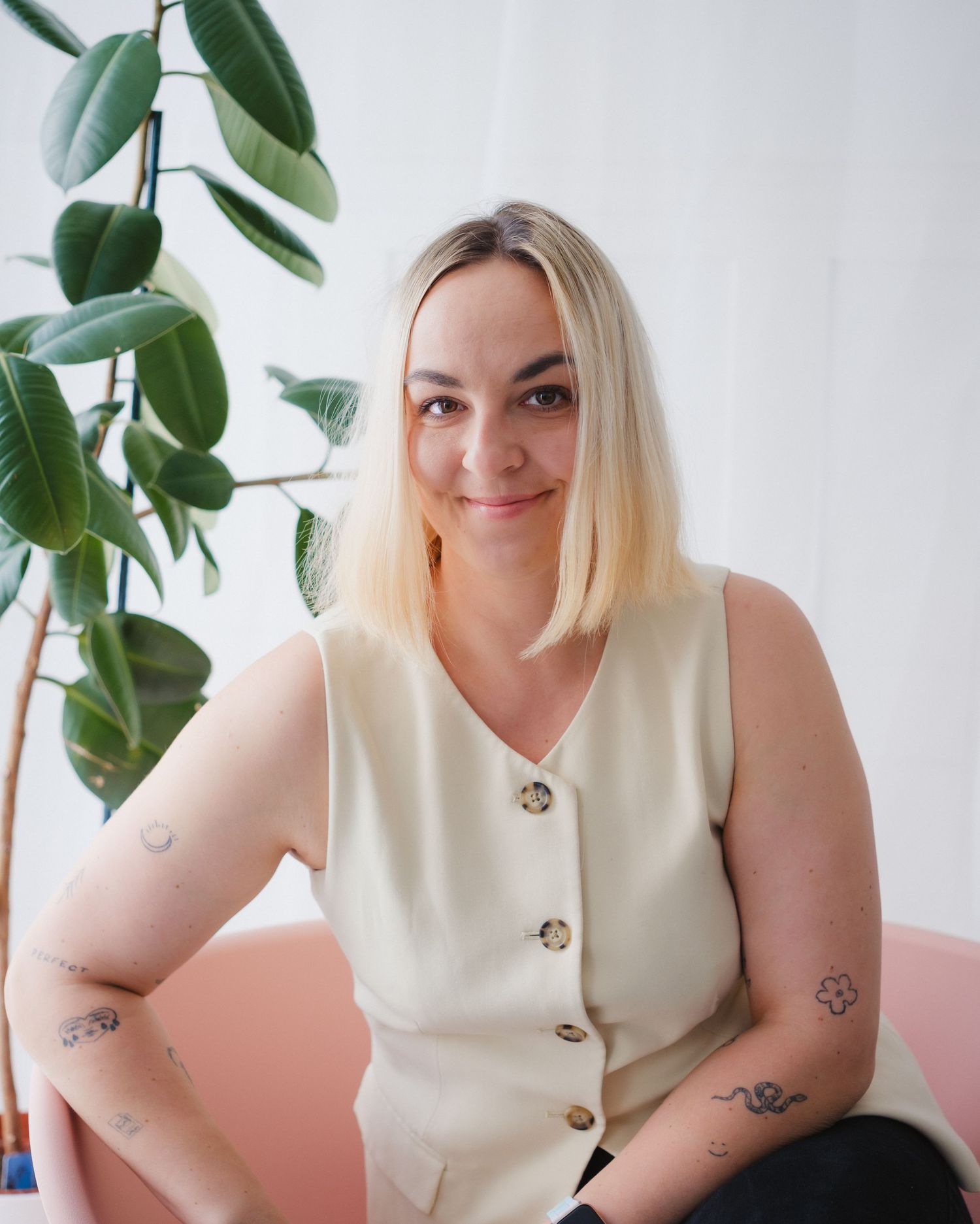 Smiling blonde tattooed woman in cream sleeveless vest seated in front of large green plant.