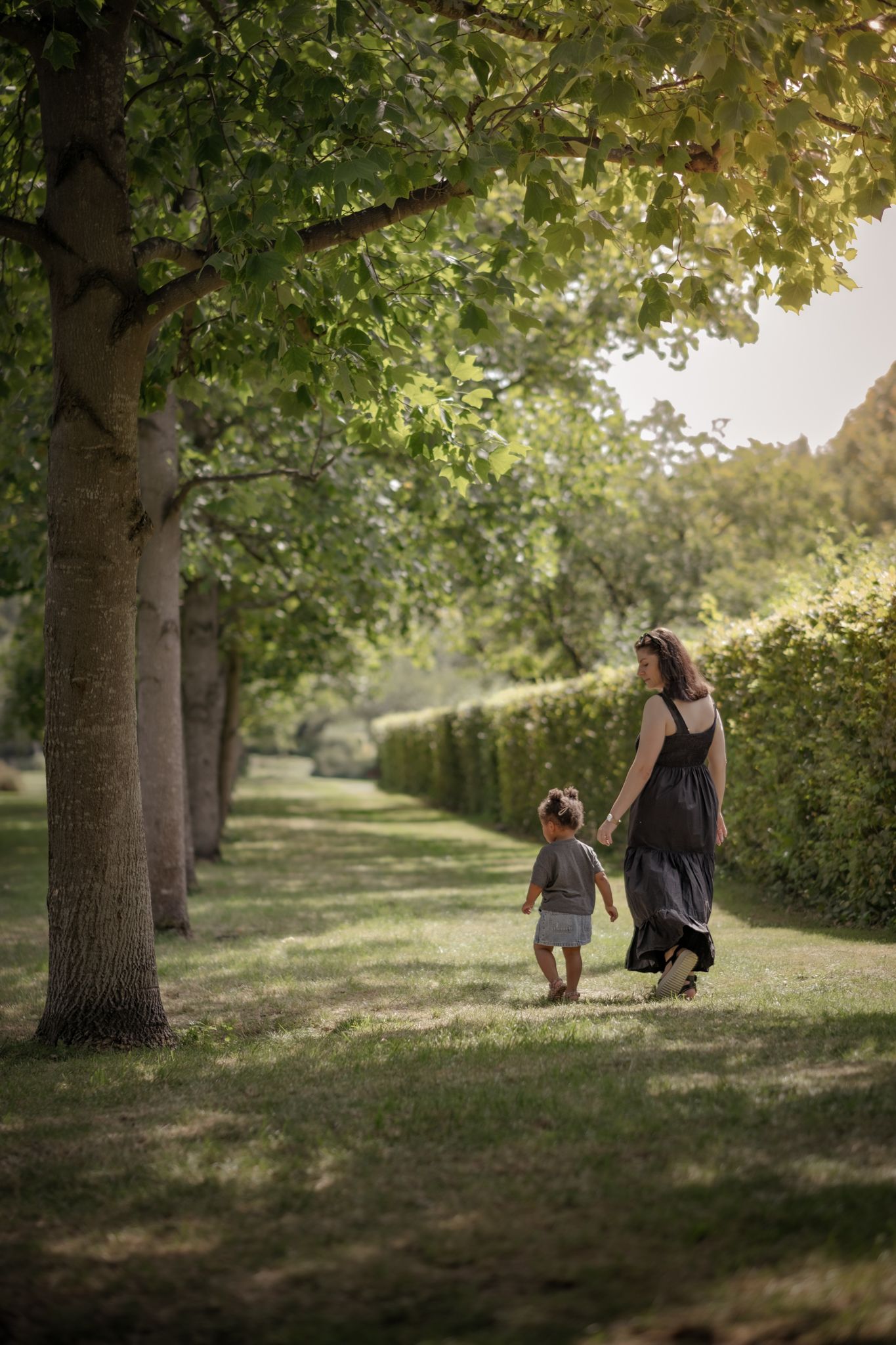 A mother and young child stroll hand in hand along a beautiful sunlit green park avenue.