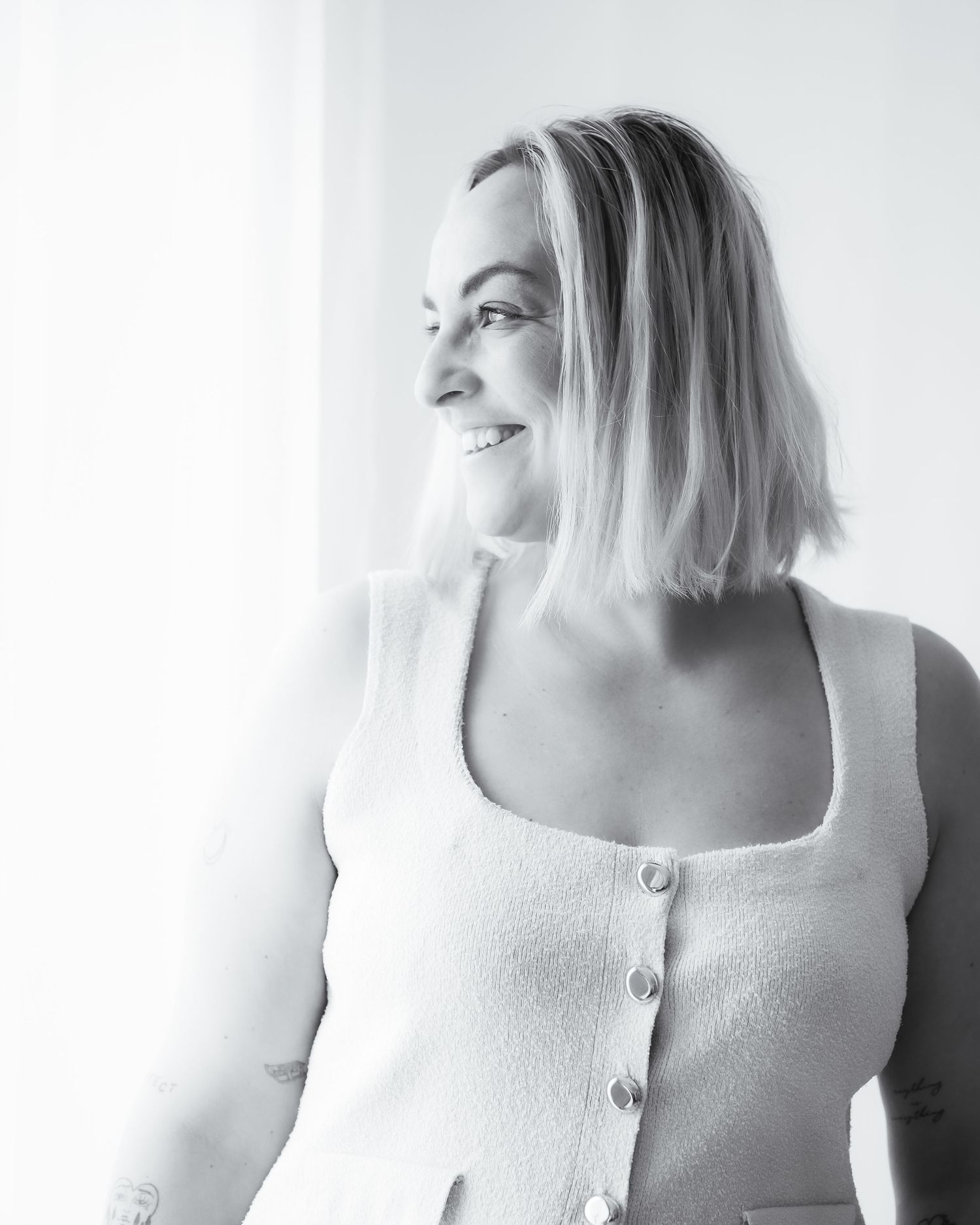 Black and white portrait of smiling blonde woman in white top by window.