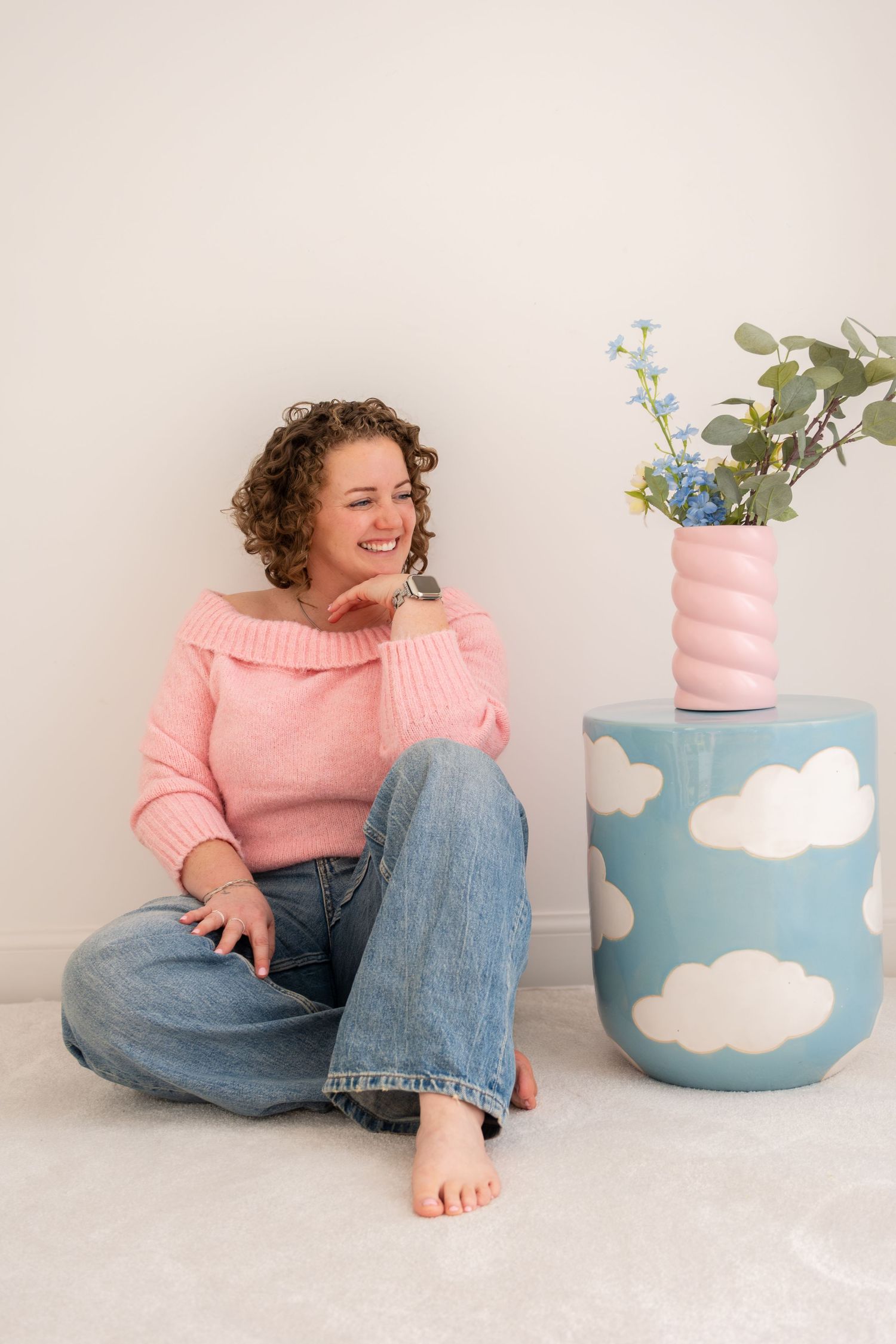 Smiling woman in pink off-shoulder sweater and jeans sitting on floor next to a blue cloud-patterned stool with pink vase.