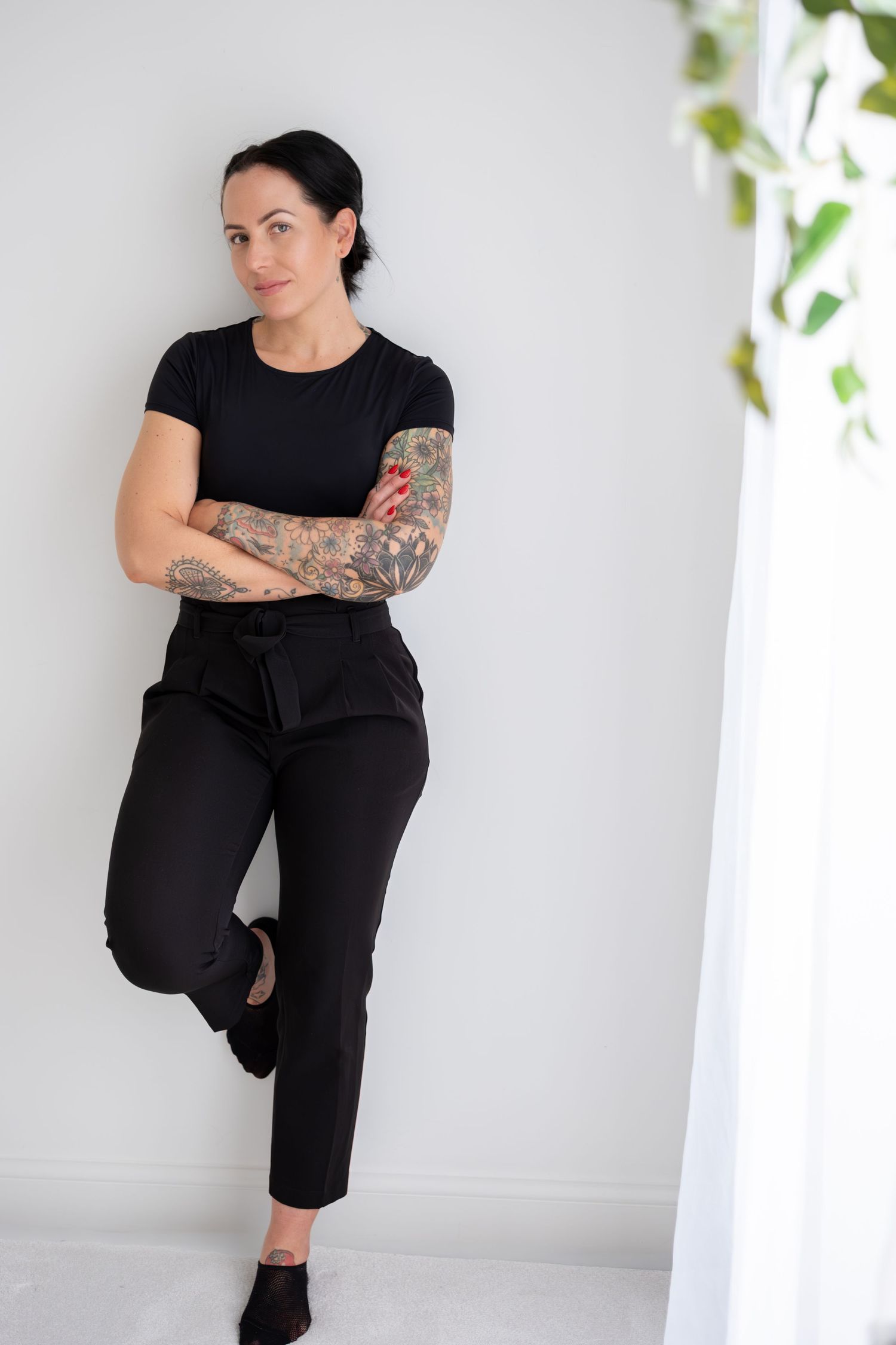 Tattooed woman in all-black outfit leans against white wall with arms crossed beside sheer curtain.