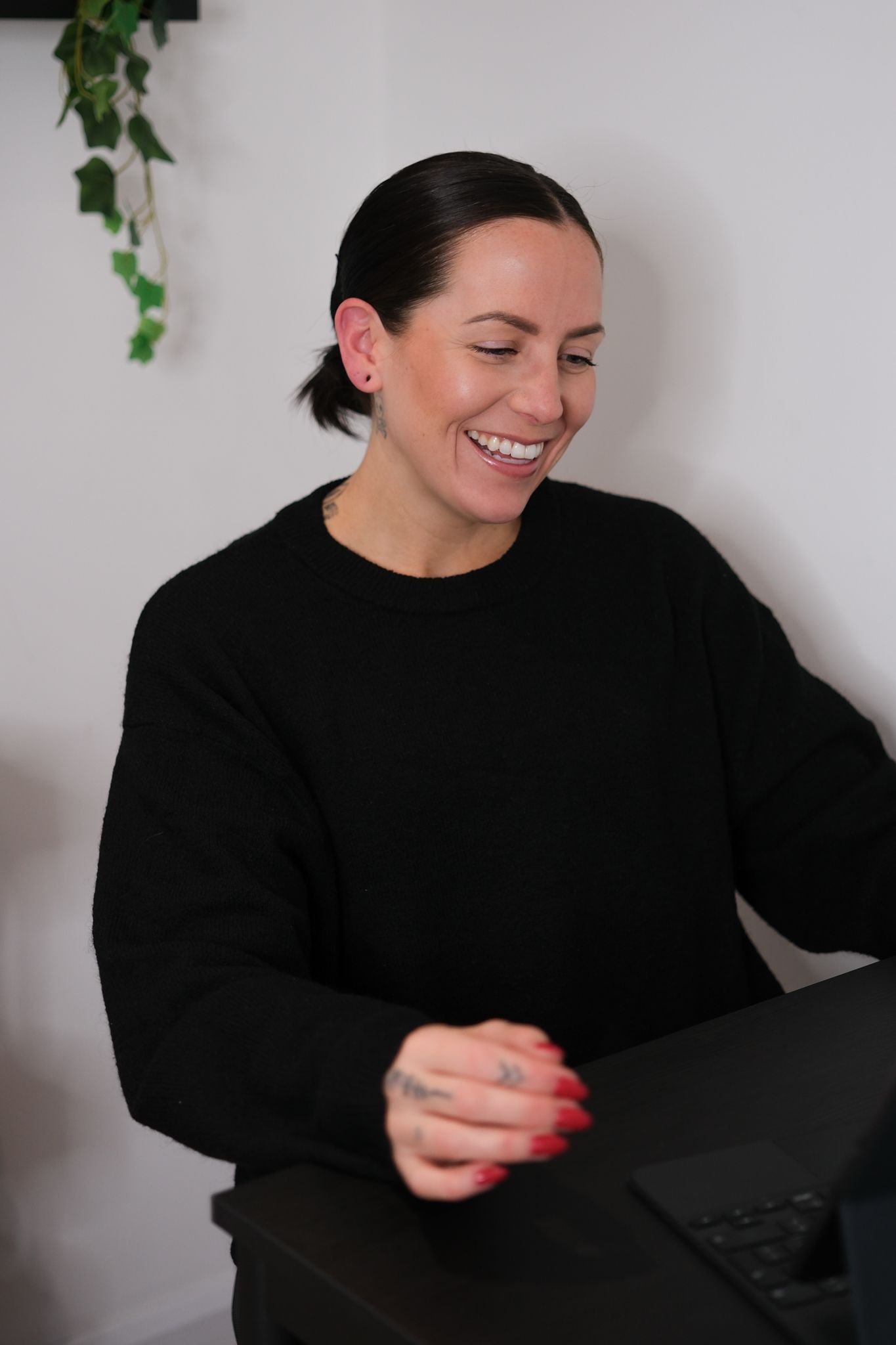 Smiling woman in black sweater laughing while working on laptop with ivy plant in background.