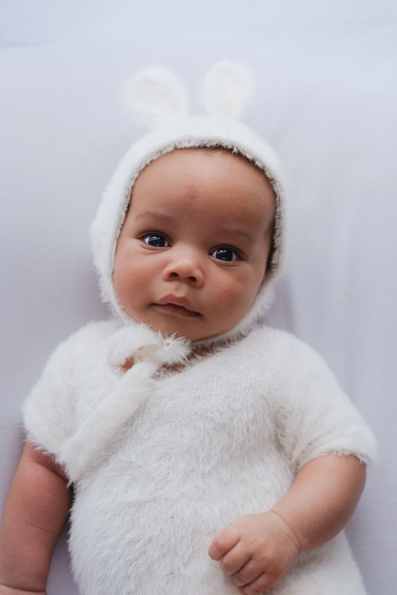 Newborn baby in white fluffy bunny costume with ears lying on light background.