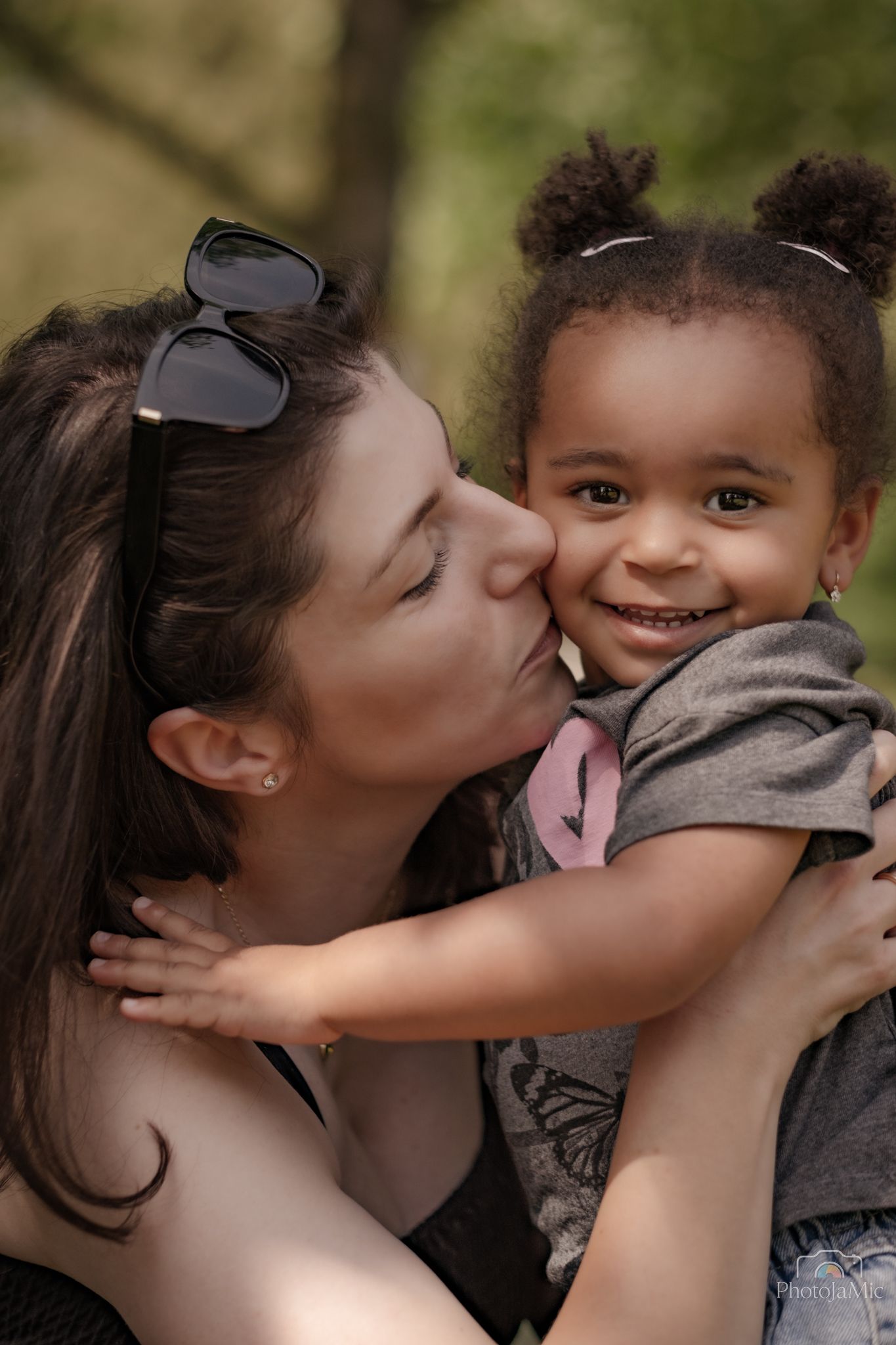 A mother lovingly kisses the cheek of her smiling young mixed-race toddler daughter outdoors.