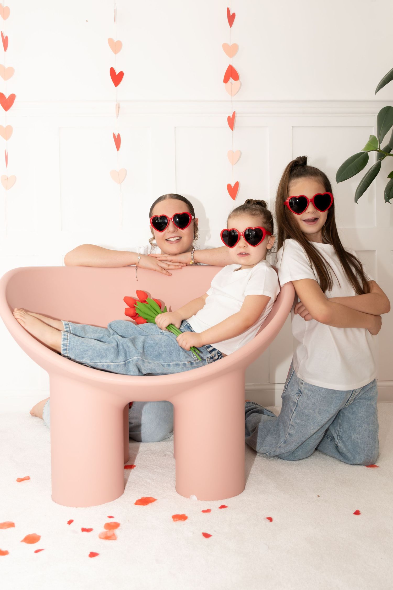 Three girls wearing heart-shaped sunglasses pose around a pink chair with Valentine's Day heart decorations.