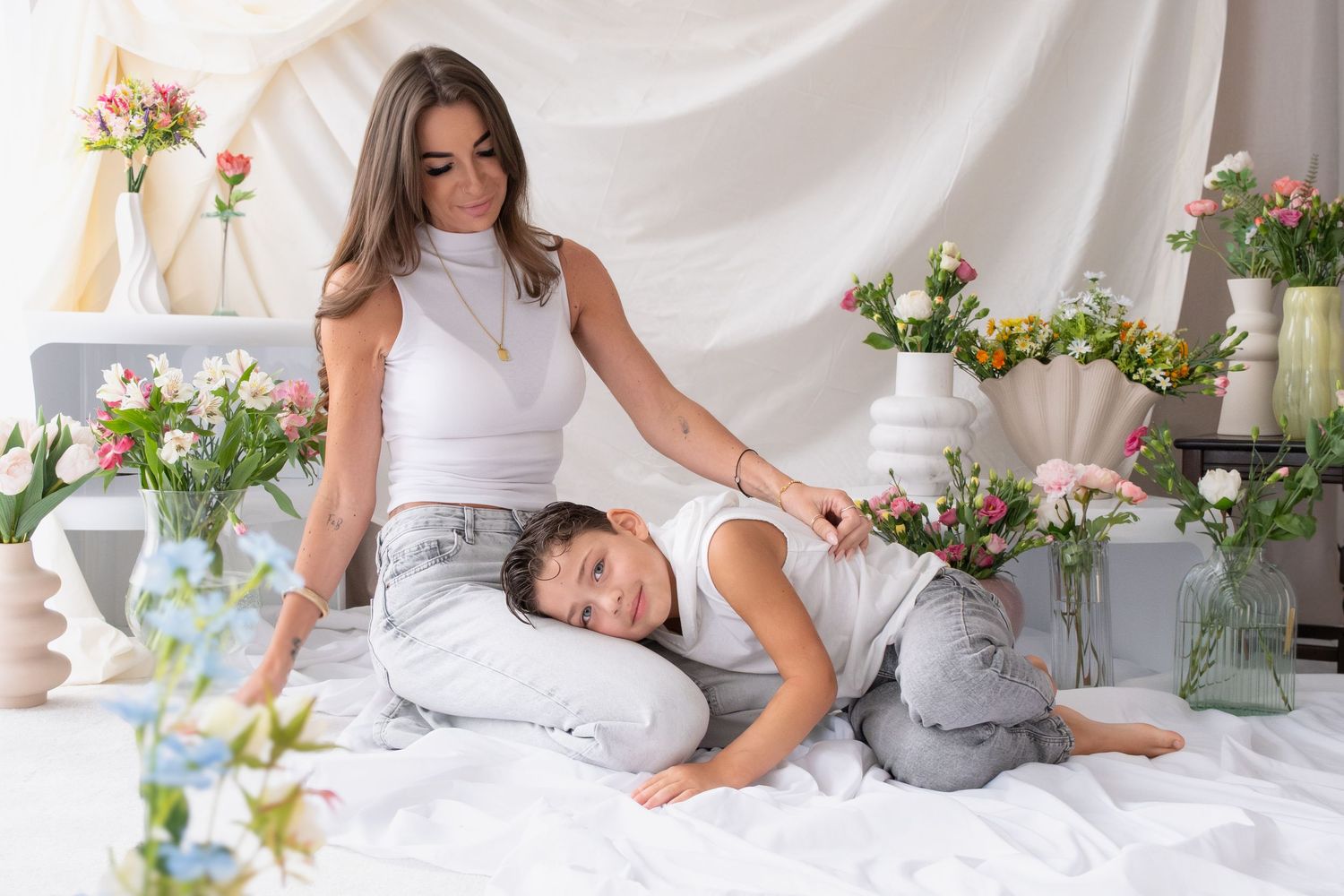 Mother and child relaxing on white bed with floral arrangements in bright airy room during photo session.