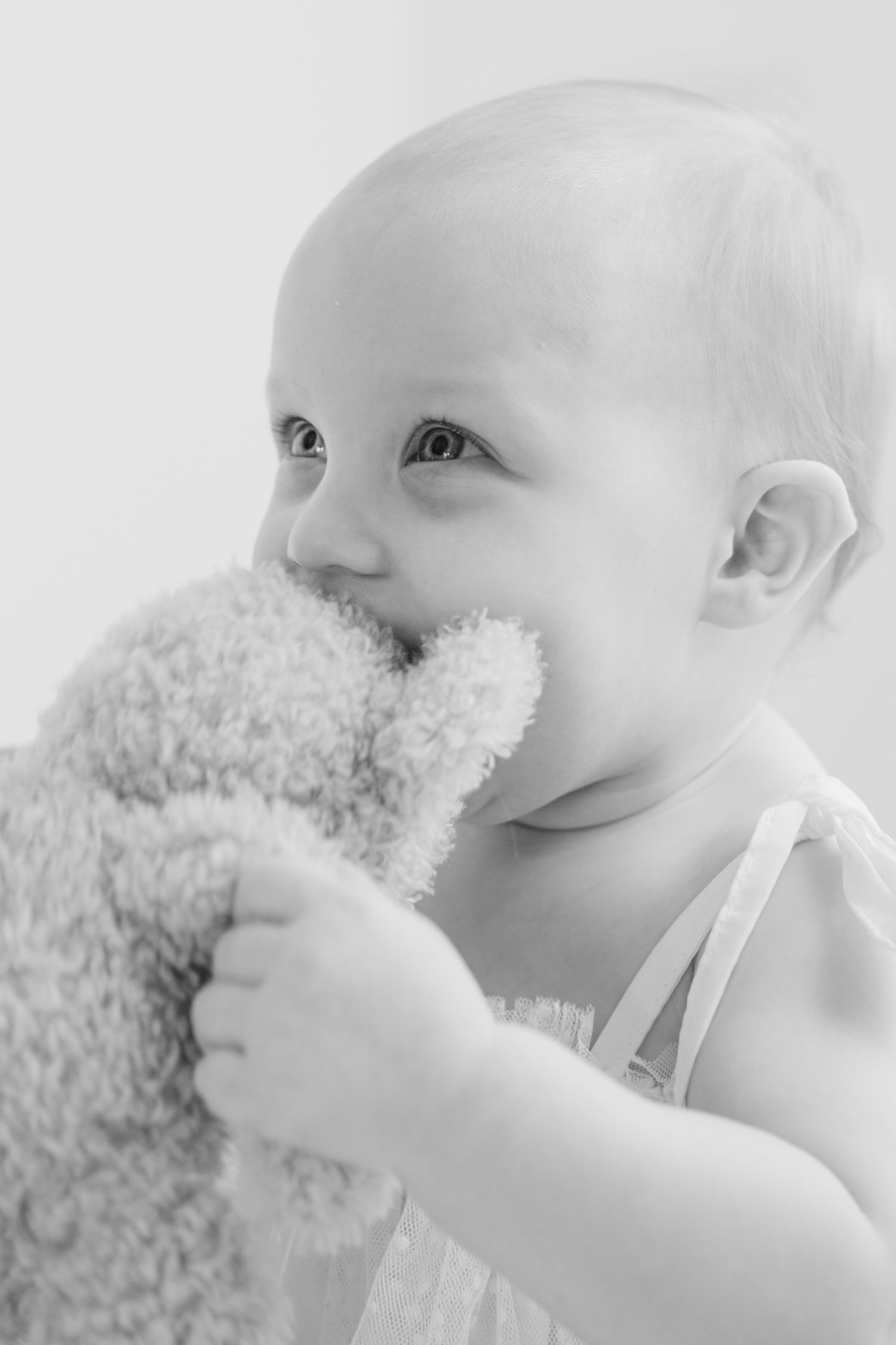 Black and white portrait of baby chewing on soft teddy bear toy.