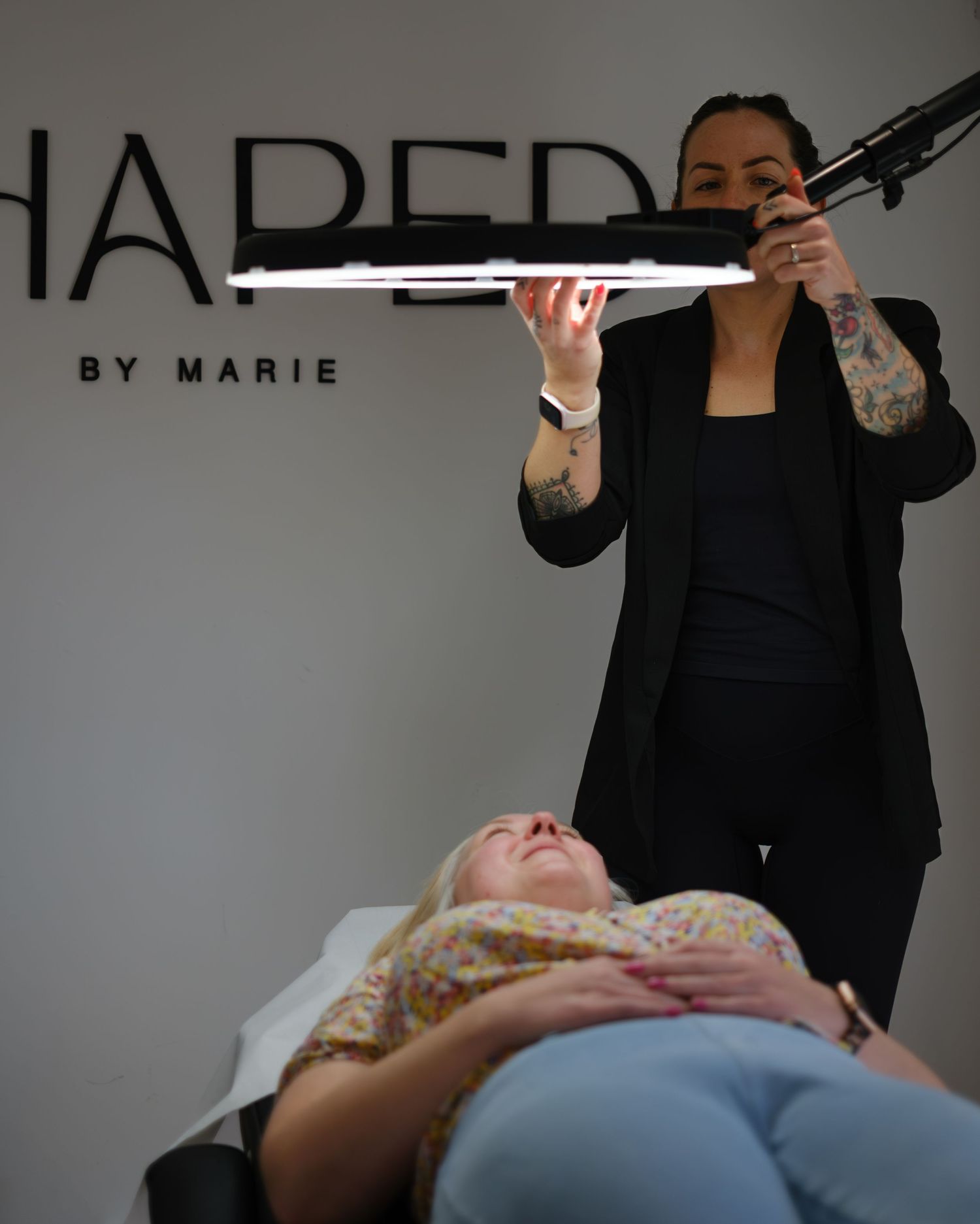 Brow artist at Shaped by Marie salon examining client with professional lighting tool.
