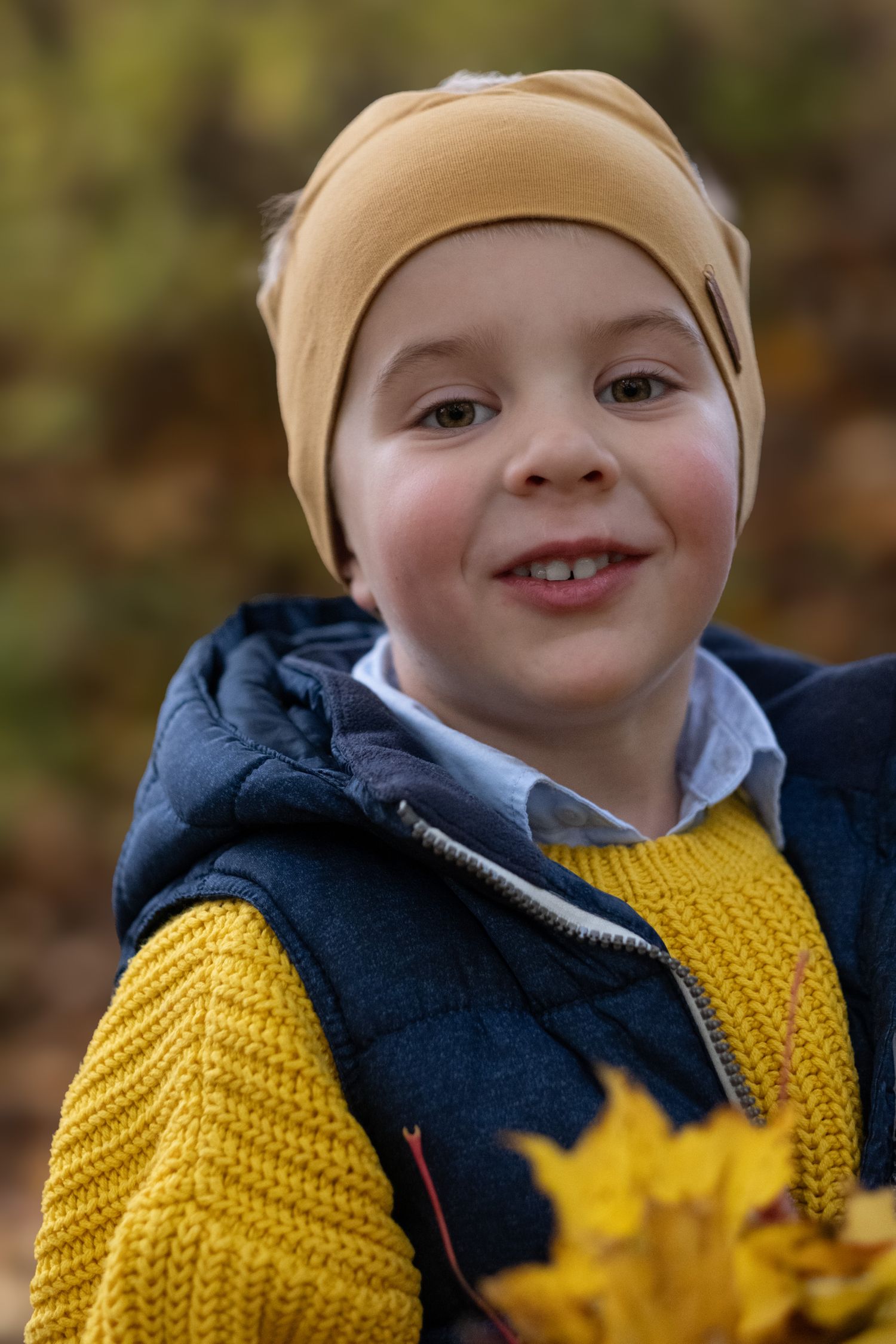Smiling young boy wearing a yellow beanie and yellow sweater holds autumn leaves in a fall forest.