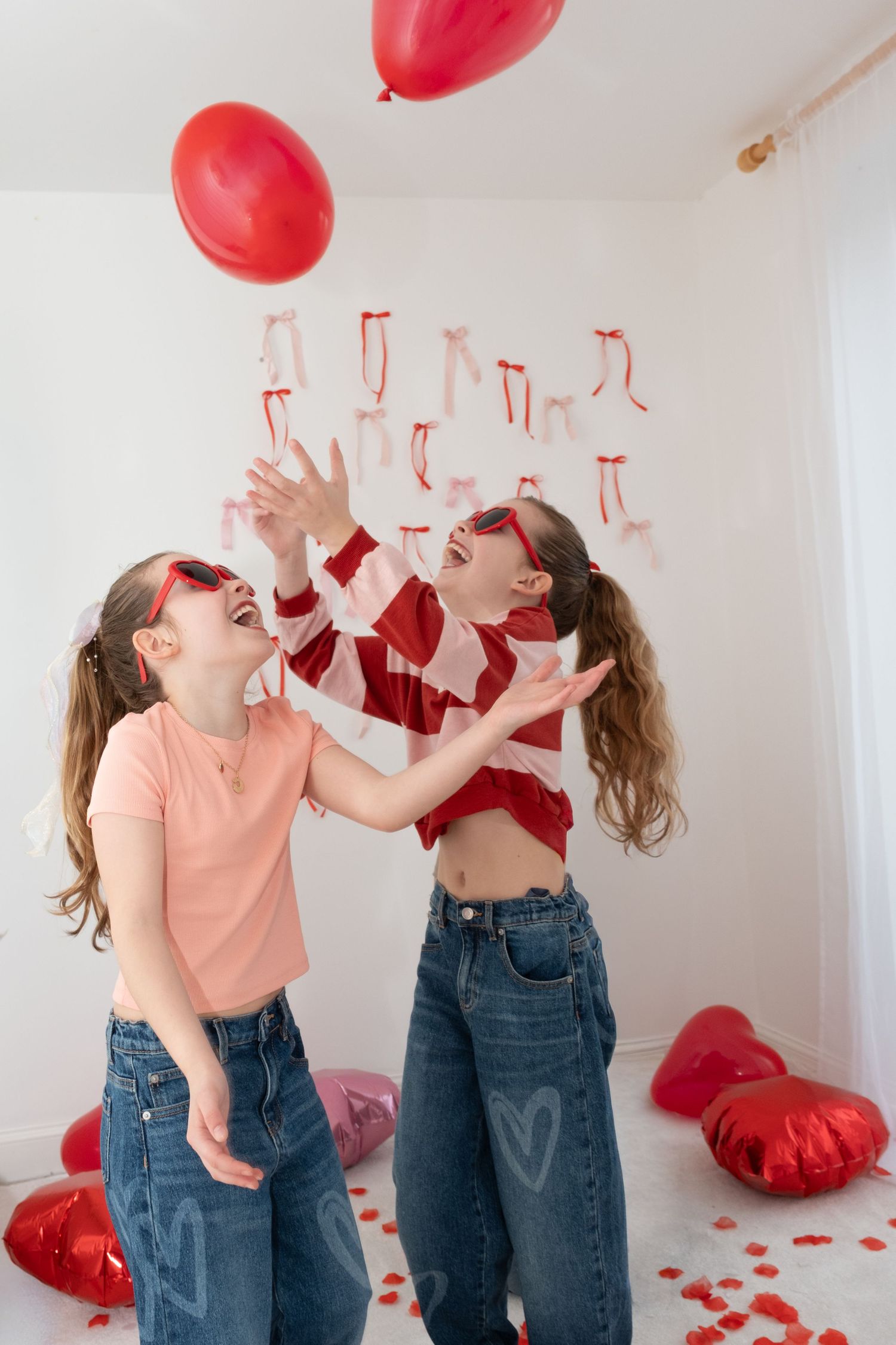 Two girls laughing and tossing red heart balloons in a Valentine's Day decorated room.