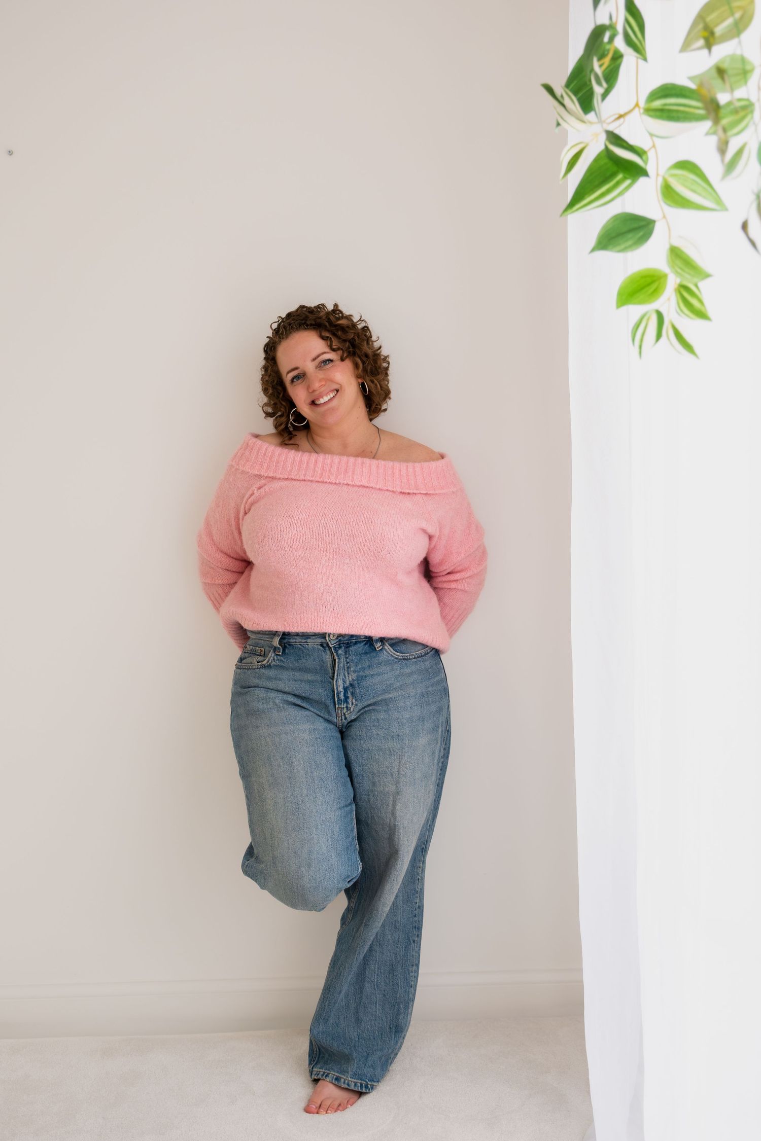 Curly-haired woman in pink off-shoulder sweater and wide-leg jeans leaning against white wall.