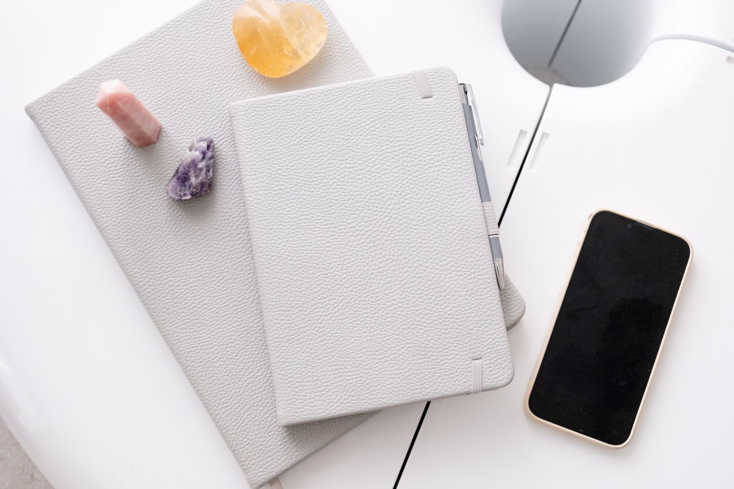Minimalist desk setup with gray notebooks, crystals, smartphone, and a white lamp on white surface.