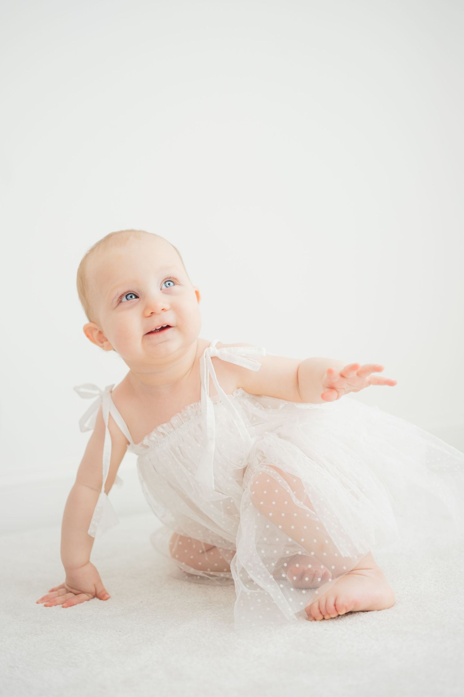 Adorable baby girl in white tulle dress crawls and reaches out on a white background.