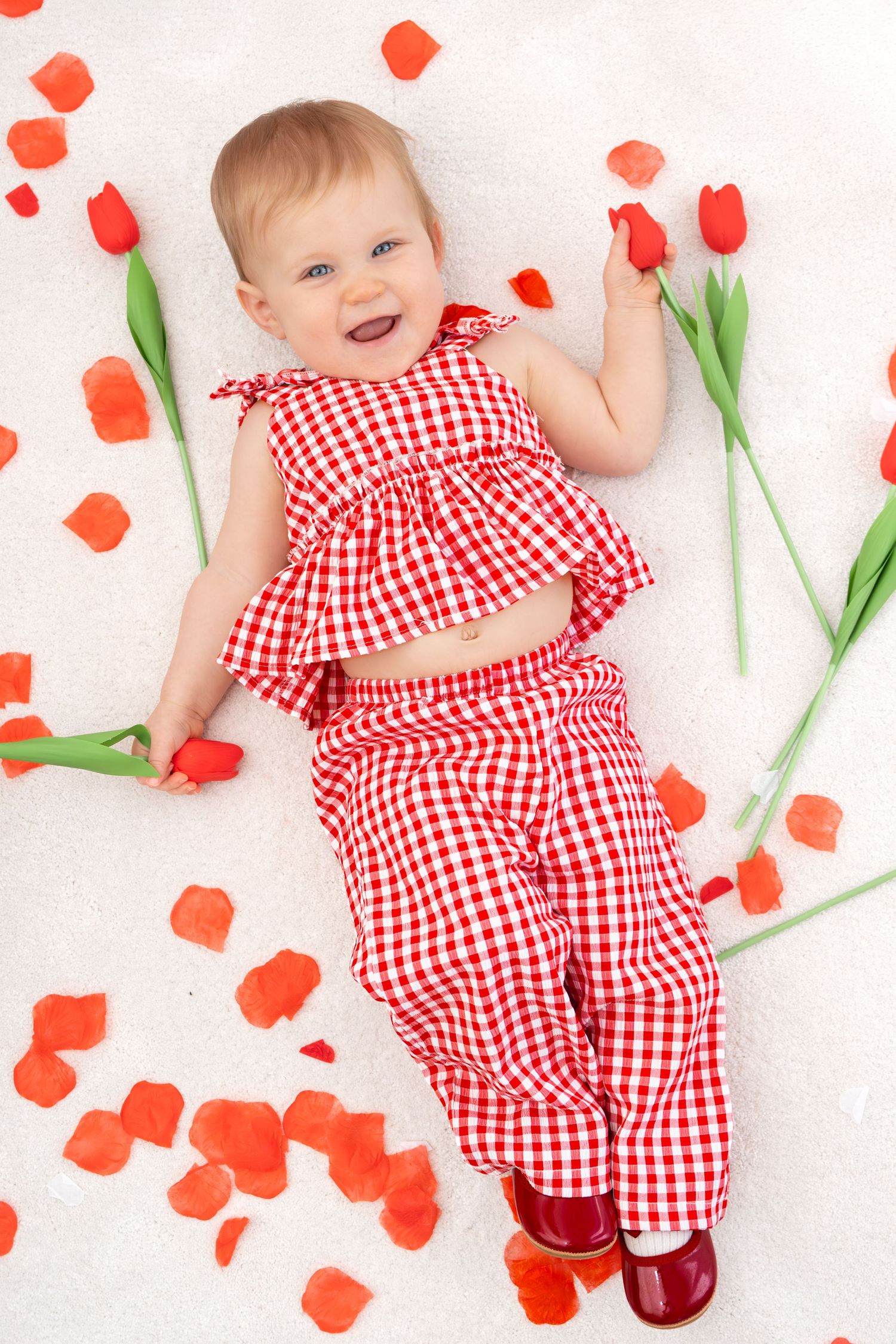 Happy baby in red gingham outfit lying among red tulips and rose petals smiling.