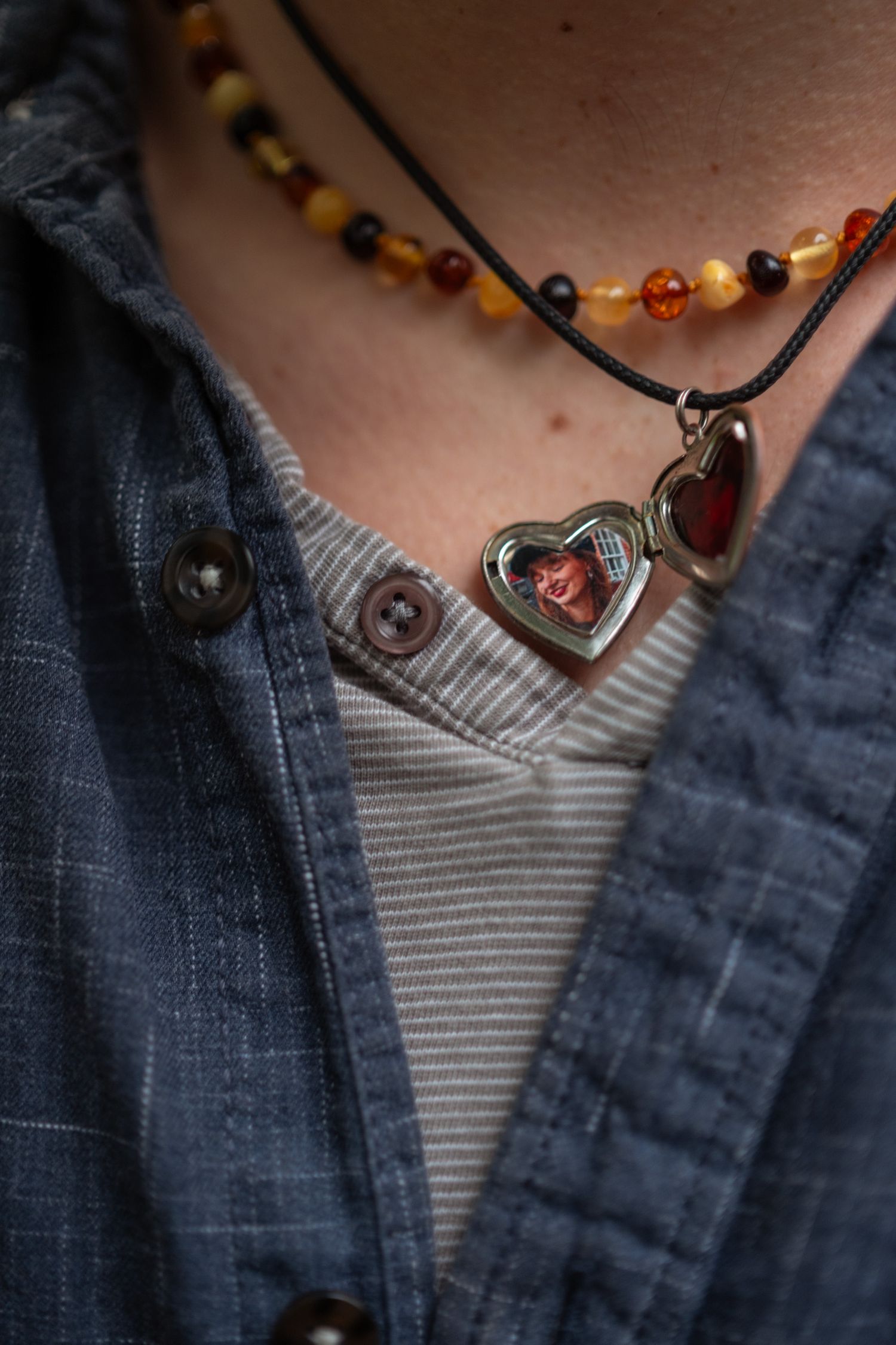 Close-up of heart-shaped locket necklace with photo pendant and amber beaded necklace layered on casual denim shirt.