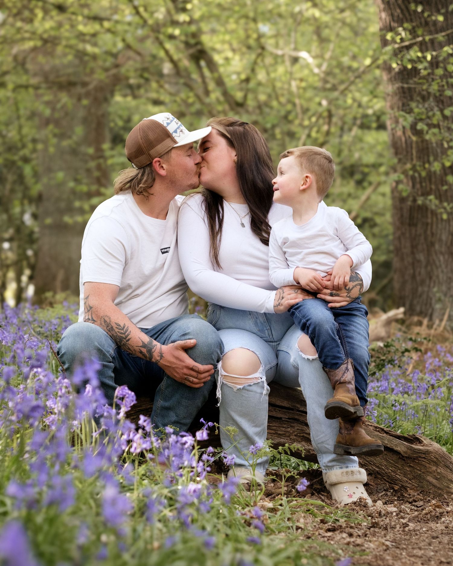 A couple kisses on a log in a bluebell forest while their toddler smiles beside them.