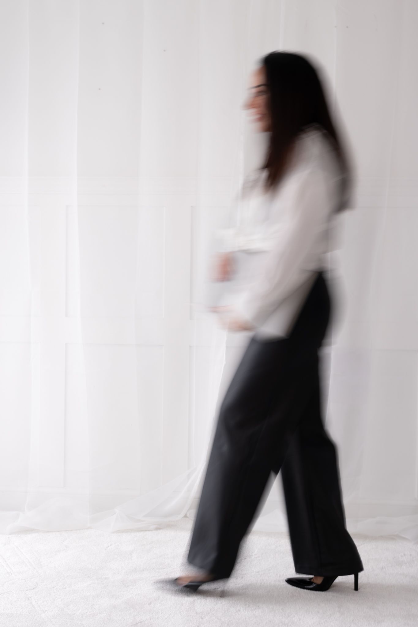 Motion blurred image of a woman in white blouse and black wide-leg pants walking past sheer white curtains.