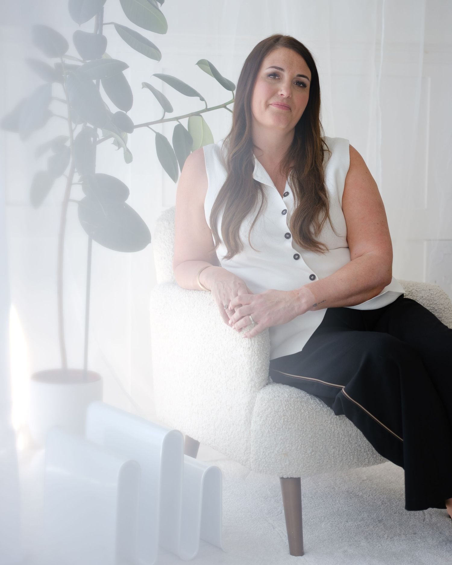 Professional woman in white sleeveless top and black pants sitting on a white chair beside a green plant.