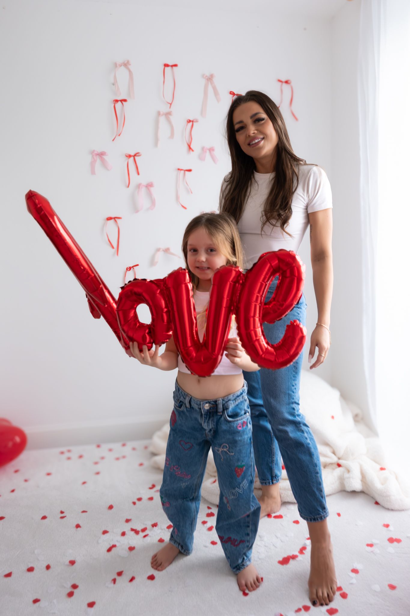 Smiling mother and daughter holding red love balloon letters in Valentine's Day decorated white room.