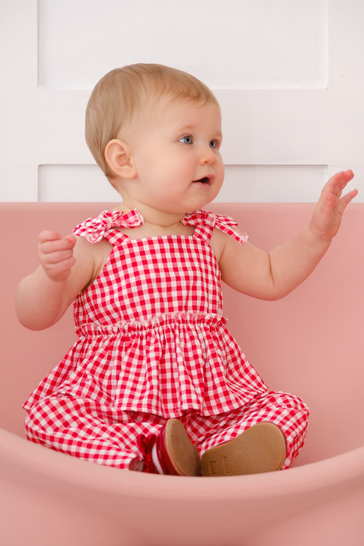 Adorable baby girl in red gingham dress sitting in a pink chair raising her hand.