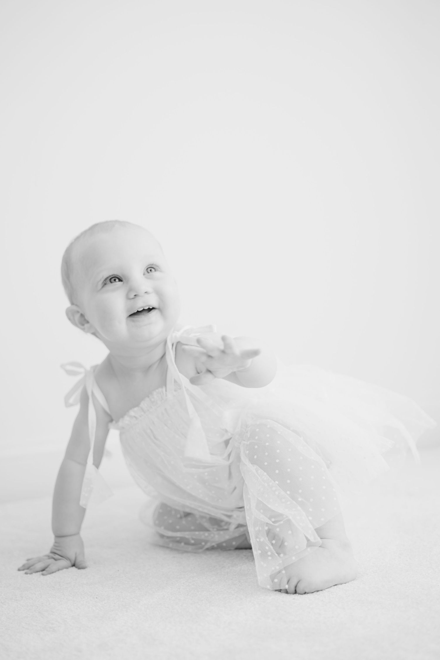 Black and white portrait of adorable baby in white lace dress crawling and looking up with joyful expression.