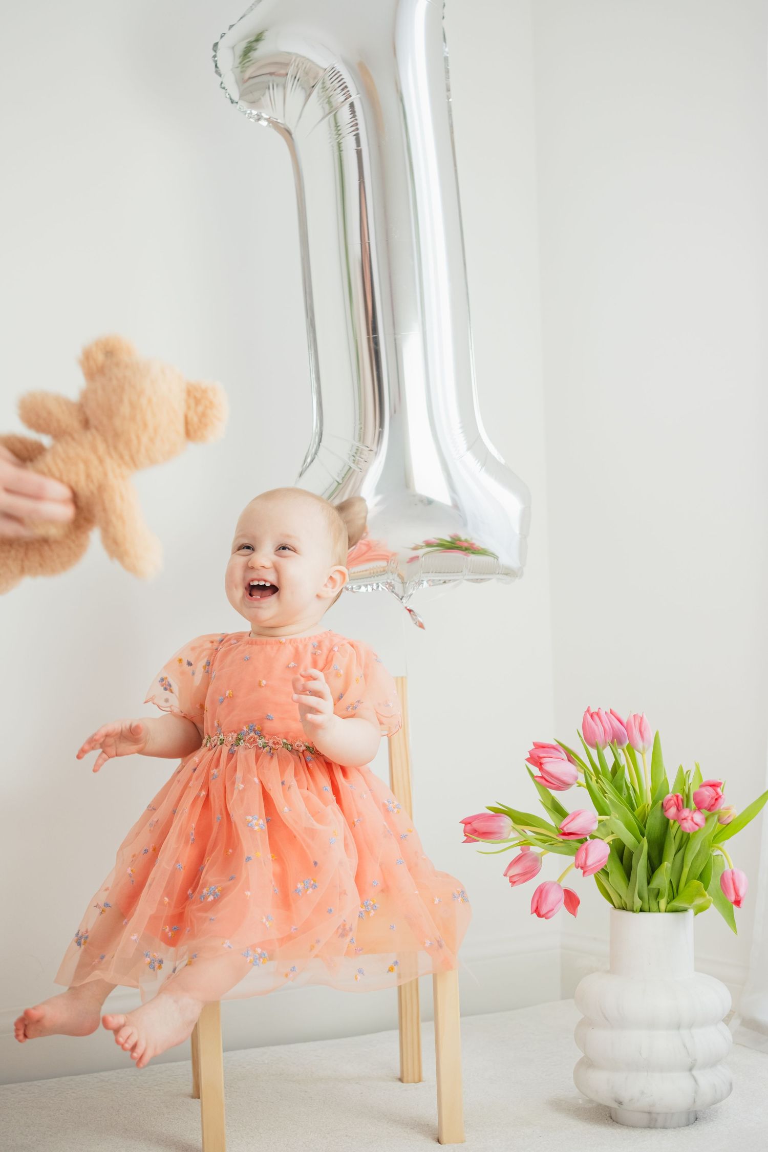 Happy baby girl in pink dress laughing on chair with number 1 balloon and tulips.