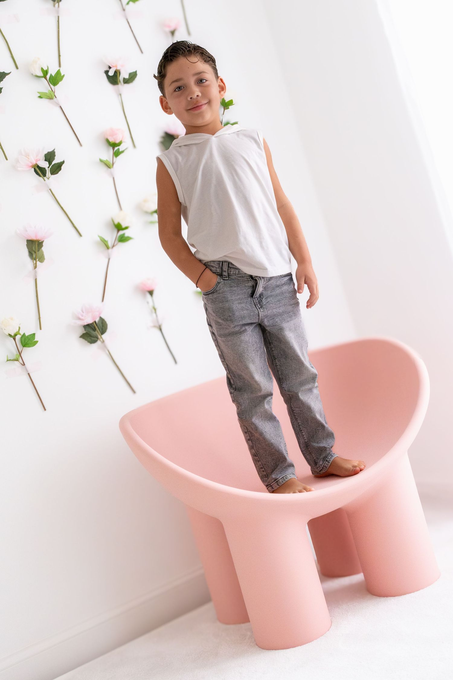 Young boy in white tank top and jeans standing on pink chair with floral wall decor.