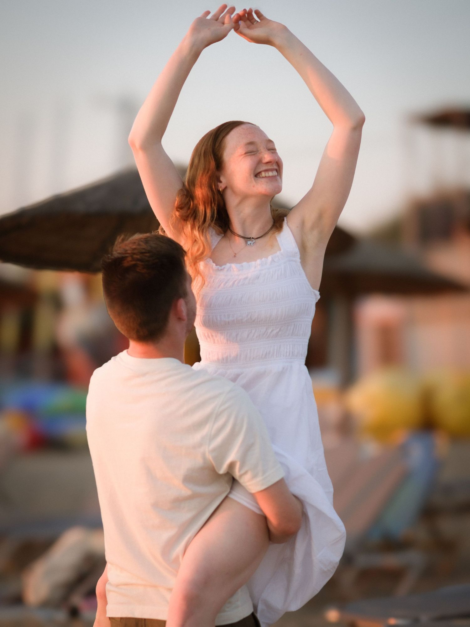 Joyful young woman in white dress lifted by her partner on a beach at golden hour sunset.