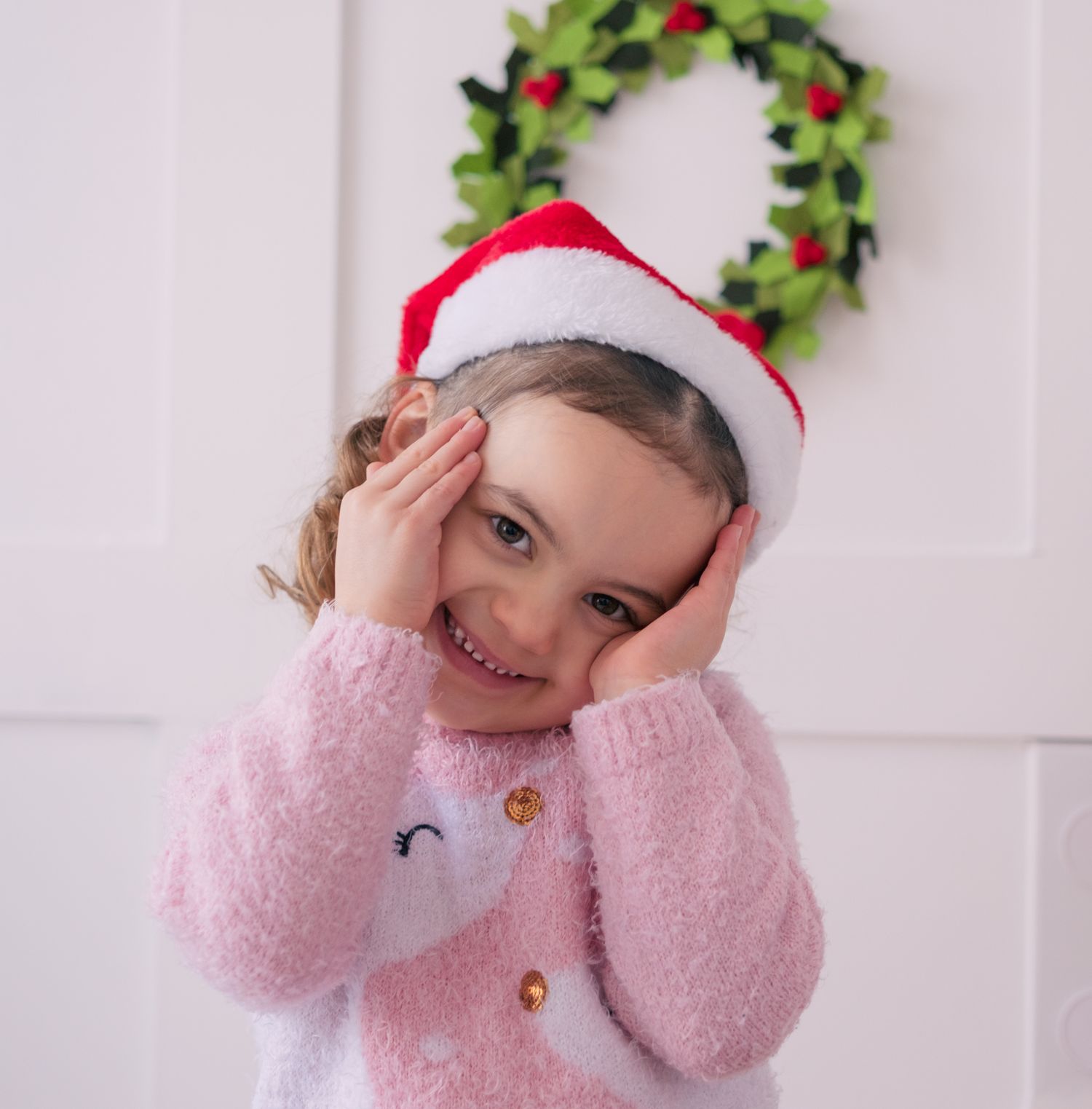 Adorable little girl in pink fuzzy sweater and Santa hat smiles with hands on cheeks near Christmas wreath.