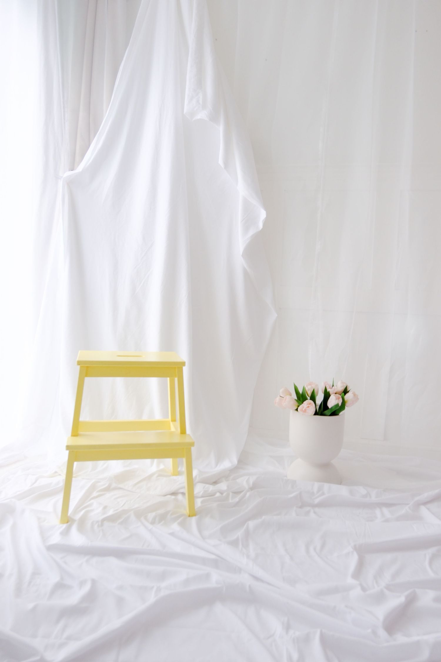 Yellow step stool beside pink tulips in white vase on white fabric background with sheer curtains.
