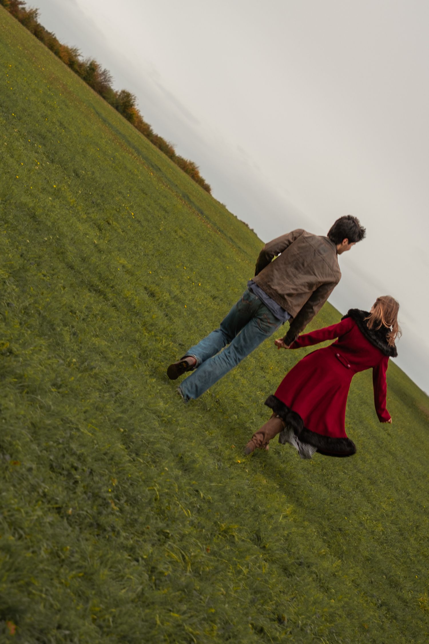 Couple holding hands running together up a green grassy hillside on an overcast day.