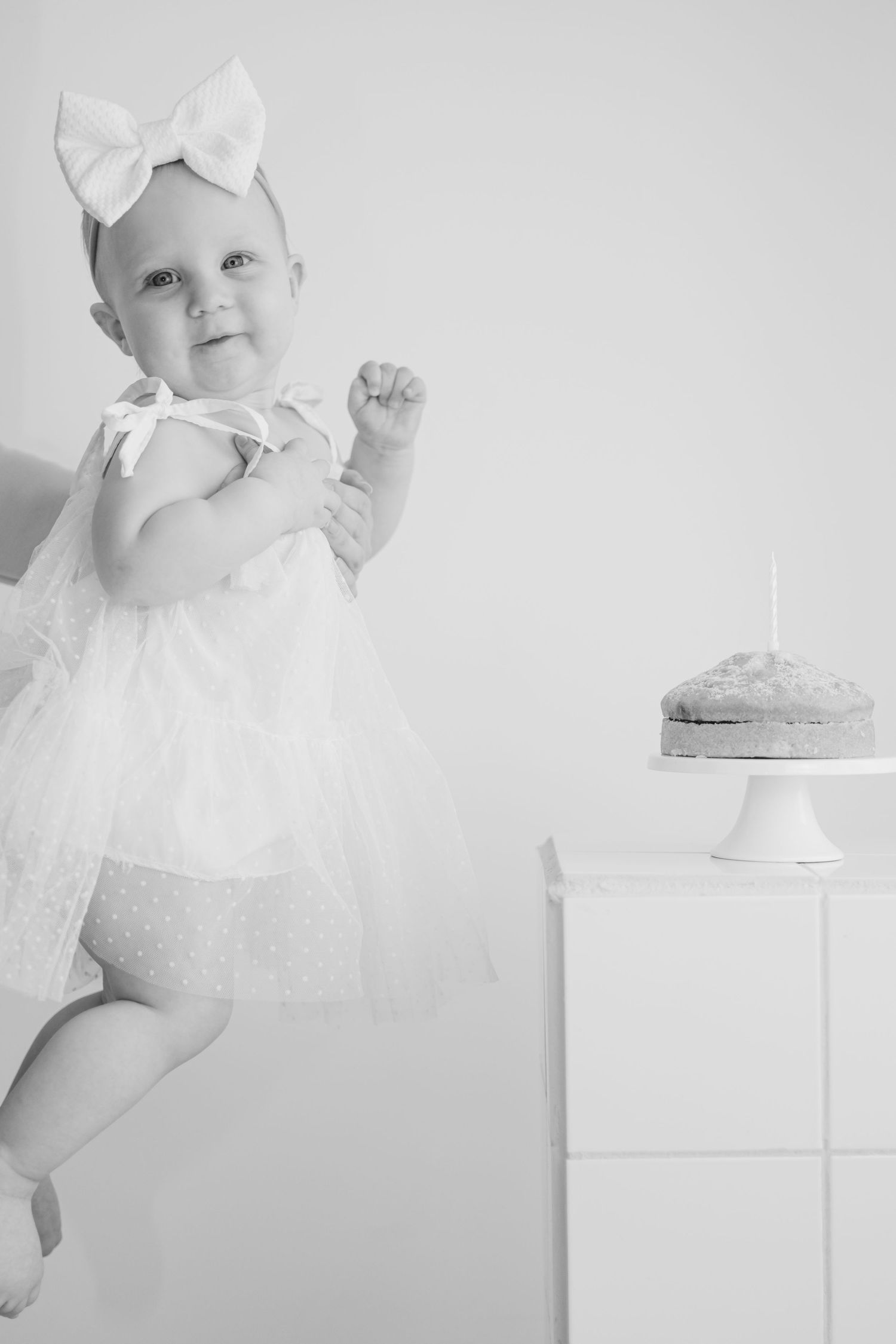 Smiling baby girl in white dress with bow headband beside birthday cake, black and white photo.