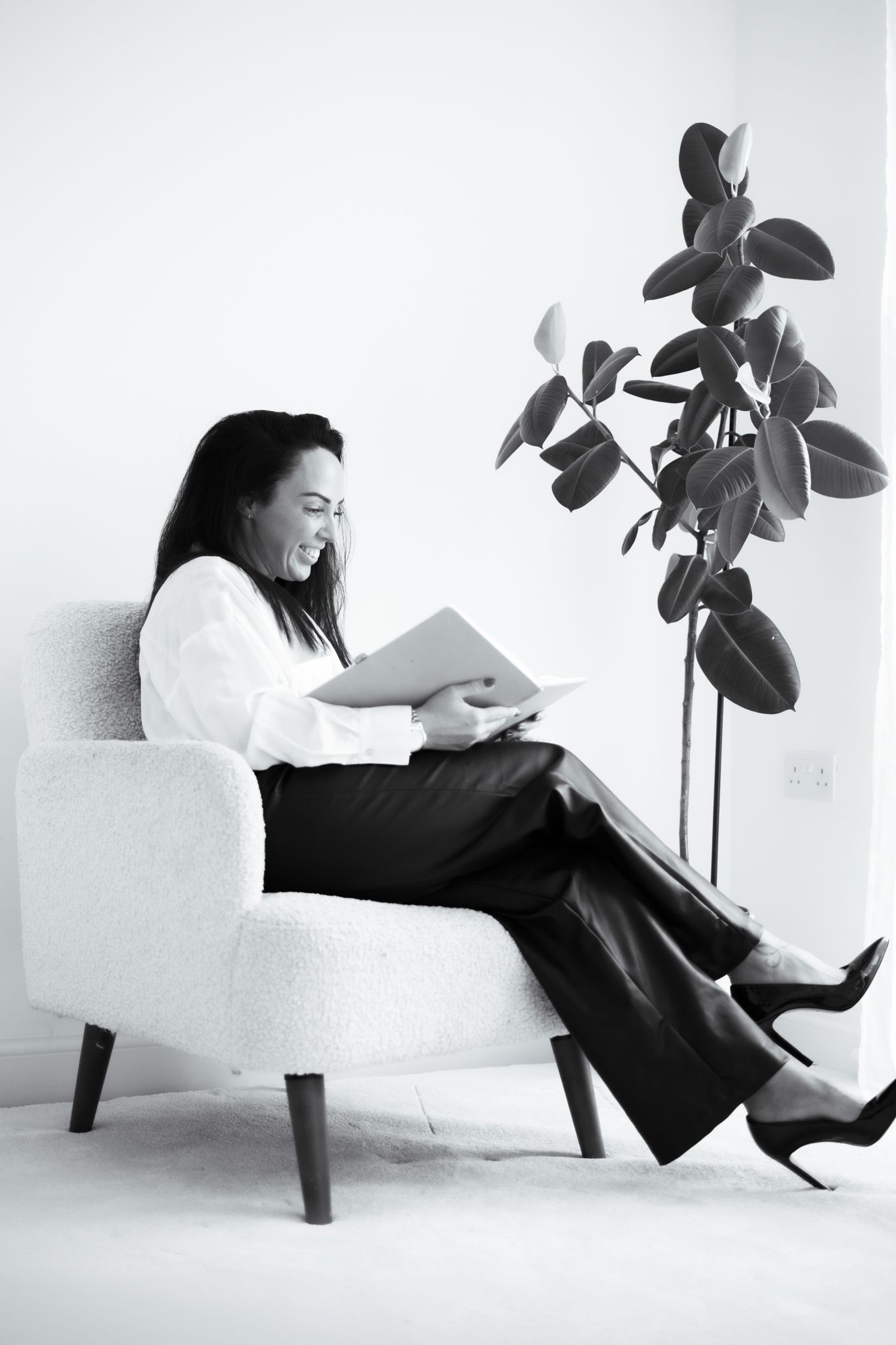 Woman in white blouse reads document while seated in white armchair beside rubber plant, monochrome.