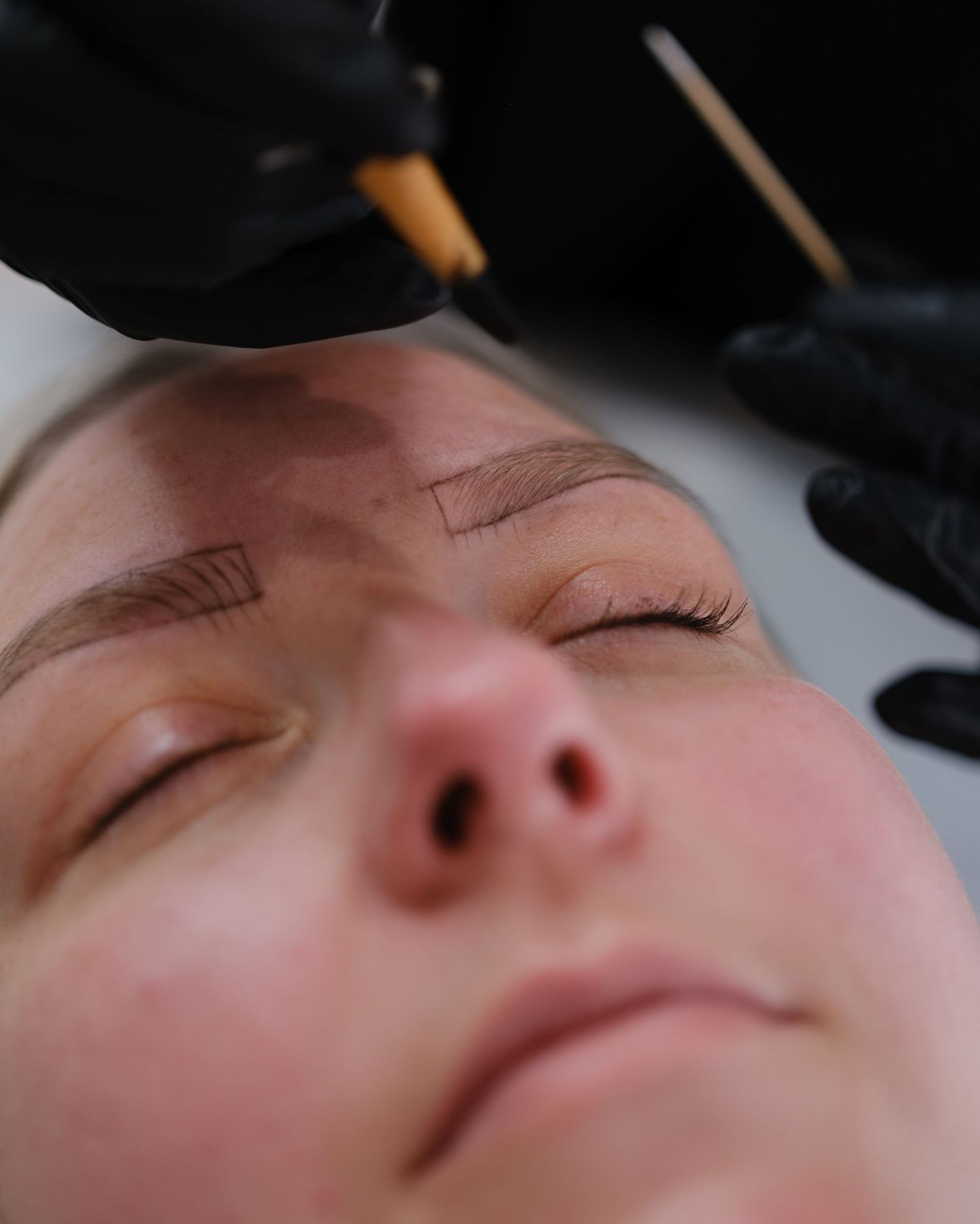Close-up of microblading eyebrow tattoo procedure with black gloved hands using a precision tool on a woman.