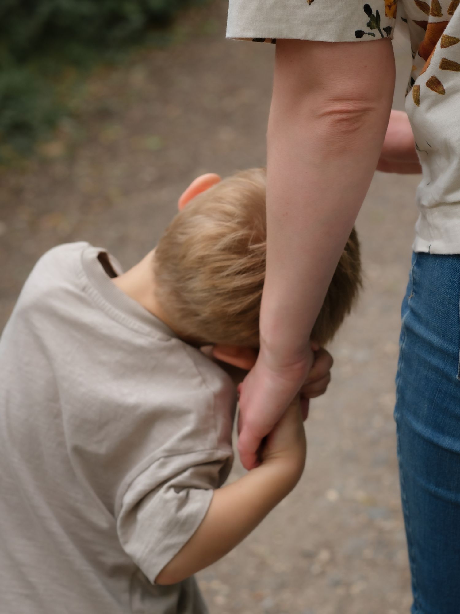 A young boy buries his face in his mother's hand while standing on a woodland path.