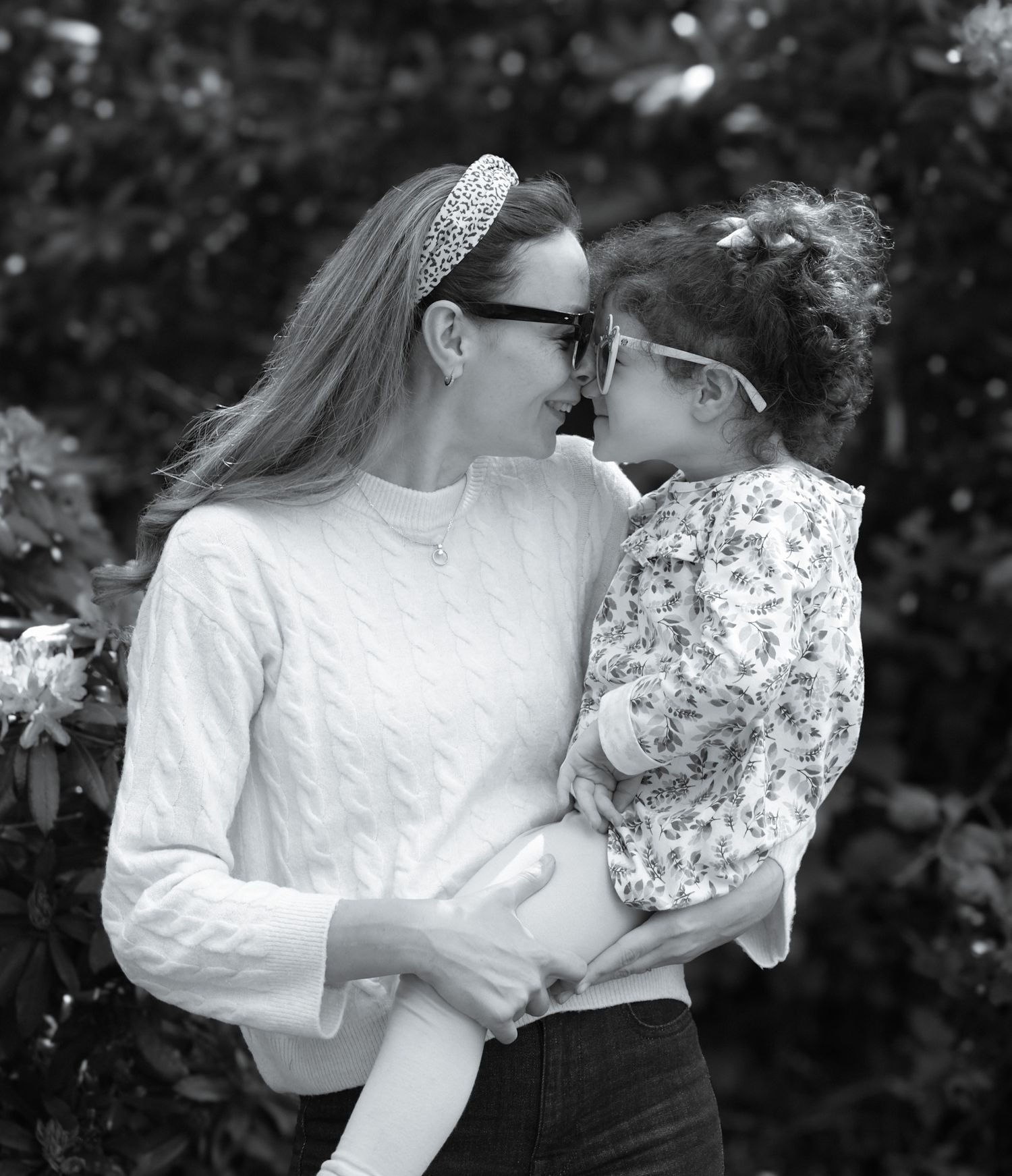 Black and white portrait of a mother and curly-haired daughter touching noses affectionately outdoors.